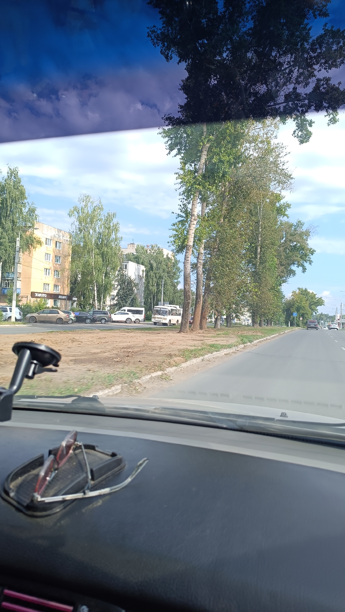 The image shows a view from inside a vehicle, looking out through the windshield. The scene is a street in Chaikovsky, Russia, on a cloudy day. In the foreground, on the dashboard, a pair of glasses rests on a non-slip mat. Outside the vehicle, to the left, are several apartment buildings with trees lining the street. Cars, including a white van and a beige sedan, are parked along the roadside. Further down the street, a white bus is visible. To the right, the road continues, with more trees and buildings in the distance. The sky is a mixture of blue and grey clouds.