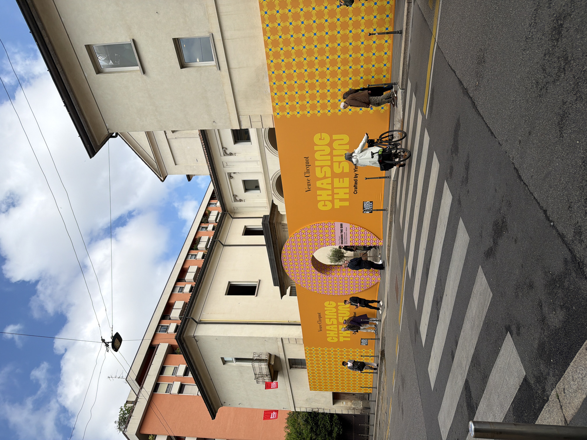 This media file captures a street scene in Milan, Italy, featuring a vibrant art installation during the Milano Design Week. The image, which is rotated 90 degrees clockwise, shows a bustling urban environment under a partly cloudy sky.
The central focus is a large, brightly colored installation spanning the length of a building wall and extending onto the sidewalk. The installation is primarily orange and yellow, featuring the text "Veuve Clicquot" and "CHASING THE SUN," along with "Crafted by Yin." The upper section of the wall installation has a repeating pattern of blue hands on a yellow background. A striking three-dimensional element, shaped like a large letter 'O' with a cutout in the middle, stands on the sidewalk, also displaying the "Veuve Clicquot" and "CHASING THE SUN" branding, along with "MILANO DESIGN WEEK" and "Opening Hours 10:00 AM - 8:00 PM." This 'O' is adorned with a pattern of pink and yellow geometric shapes.
Several people are present in the scene. One individual is seen riding a bicycle across a prominent zebra-striped pedestrian crossing. Others are walking or standing near the large installation, possibly engaging with the display. The street itself is asphalt, with painted white pedestrian stripes and a yellow line visible.
In the background, various buildings line the street, exhibiting a mix of architectural styles, including older structures with windows and balconies, and a more modern building with multiple windows and terraced sections. Overhead, streetlights and numerous electrical wires crisscross the blue sky dotted with white clouds. The overall impression is a lively daytime setting, showcasing a public art or promotional event.