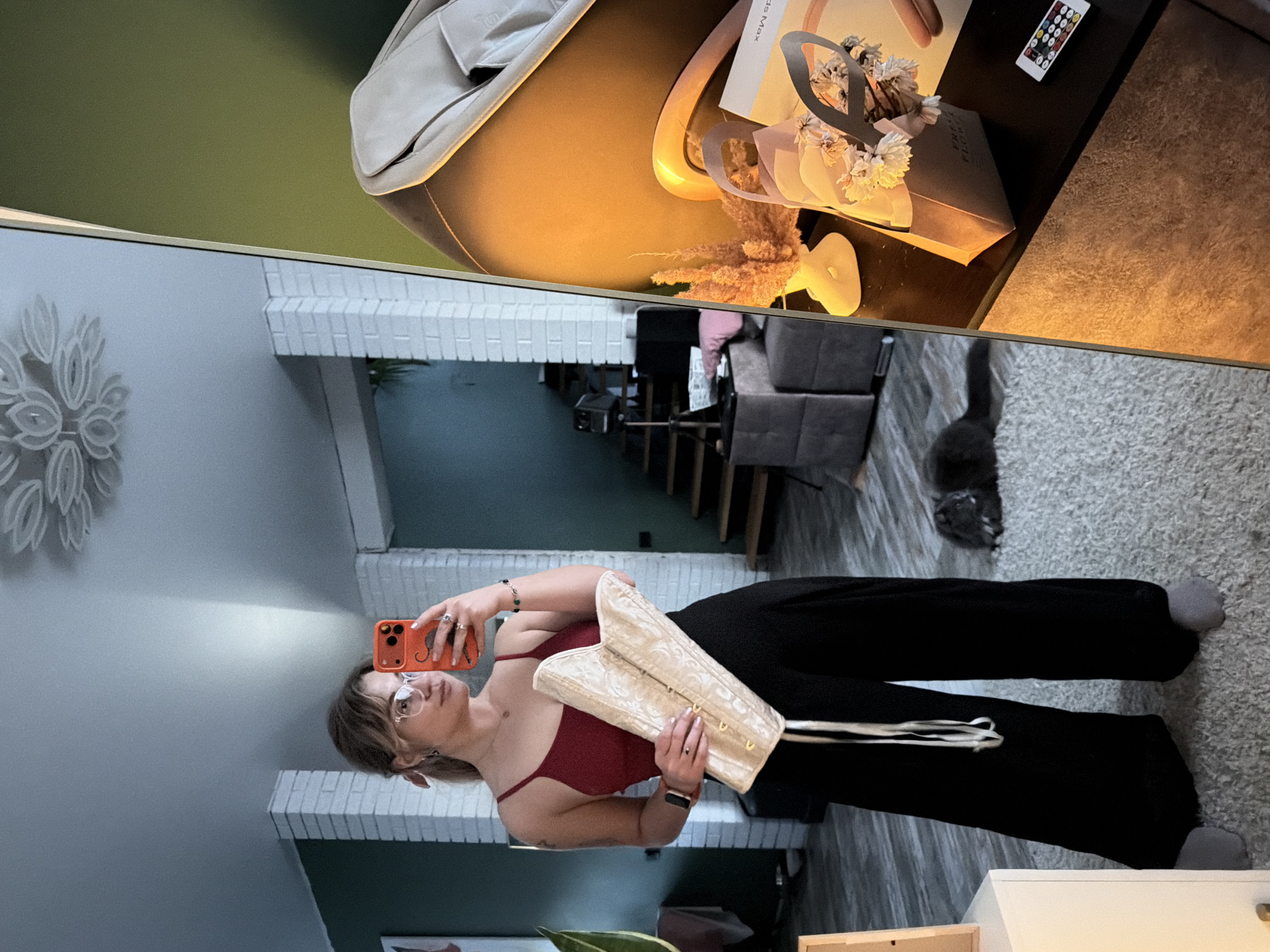A woman is reflected in a mirror, holding a corset and an orange cell phone. She has blonde hair and is wearing glasses, a red spaghetti-strap top, and black pants. She is standing in a room with white brick detailing, a green wall, a decorative flower on the wall, and a glimpse of stairs leading to another floor. In the mirror's reflection, there is a grey sofa, a black cat, and a side table with a lamp, remote control, and vase of flowers. The location may be Большая Шильна, Russia.