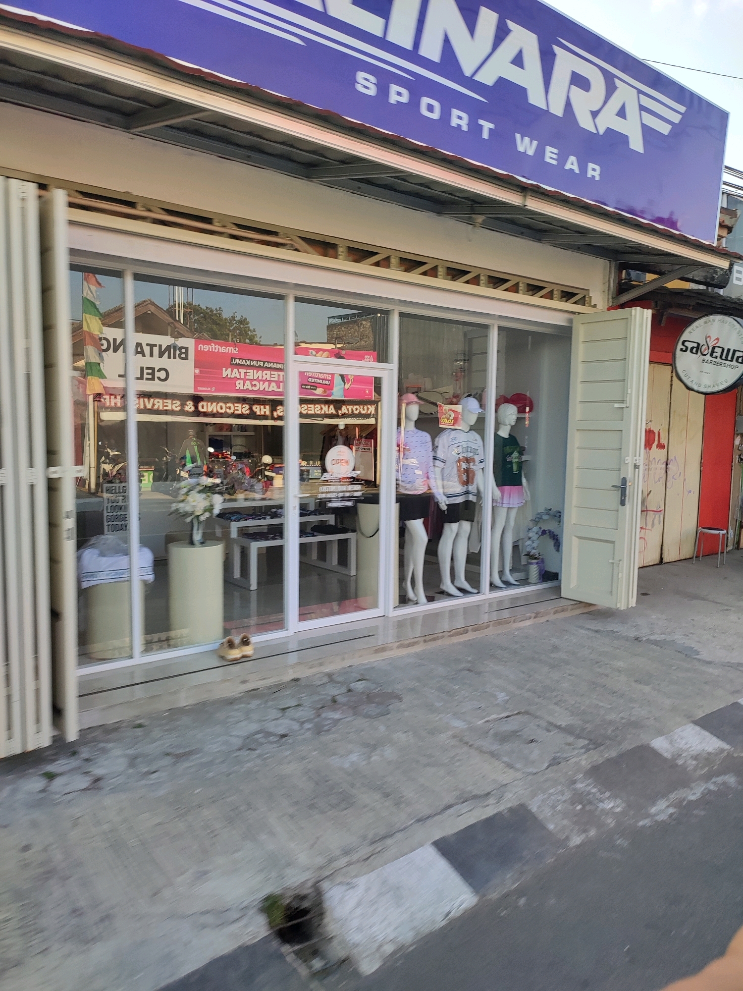 The image shows a storefront in Magelang, Indonesia, featuring a sign that reads "SINARAF SPORT WEAR". The store has large glass windows displaying mannequins dressed in sportswear. Inside, there are shelves with clothing and a vase of flowers. To the right of the storefront, there is a red building with a round sign that says "SADEWA BARBERSHOP". The street in front of the store is paved with concrete and has a zebra crossing. The overall impression is that of a commercial area on a street in the city.