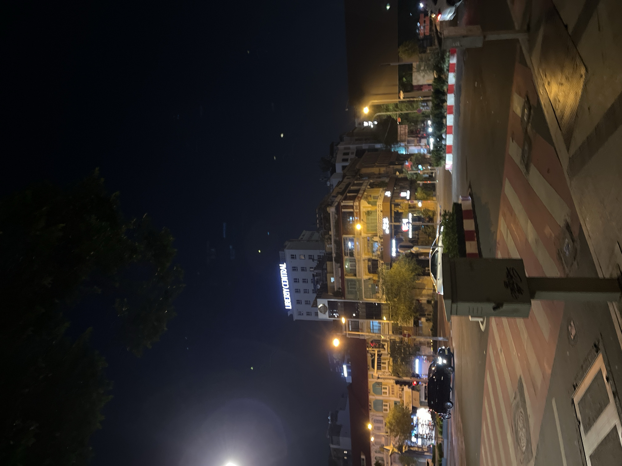 This is a night-time shot of a street in Thủ Đức, Vietnam. The "Liberty Central" building is visible in the background with its illuminated sign. Streetlights and neon signs provide illumination. A traffic signal is positioned near the camera, and a crosswalk is painted on the street. Trees line the street. Several cars and motorcycles are moving along the street. The sky is dark.