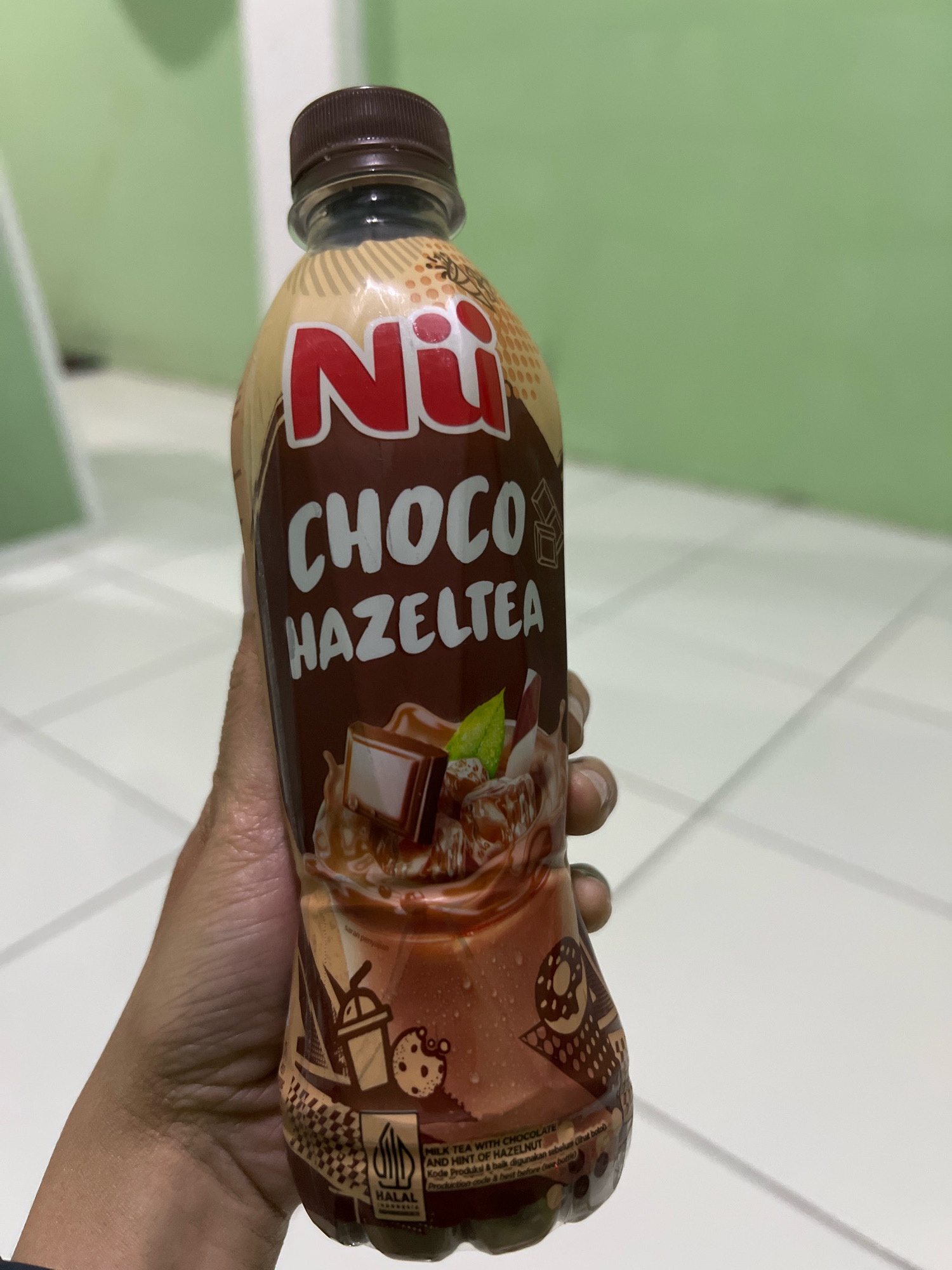 A person's hand is holding a brown plastic bottle of "Nii Choco Hazeltea" drink. The bottle has a brown cap and a light brown label with red lettering for the brand name "Nii" and white lettering for "Choco Hazeltea". The label features illustrations of chocolate, hazelnuts, and a cookie. The background is a light green wall and a tiled floor. The image is taken from a medium close-up perspective, with the bottle slightly tilted. No other specific details about the location in Tlogoadi, Indonesia can be determined from the provided image.
