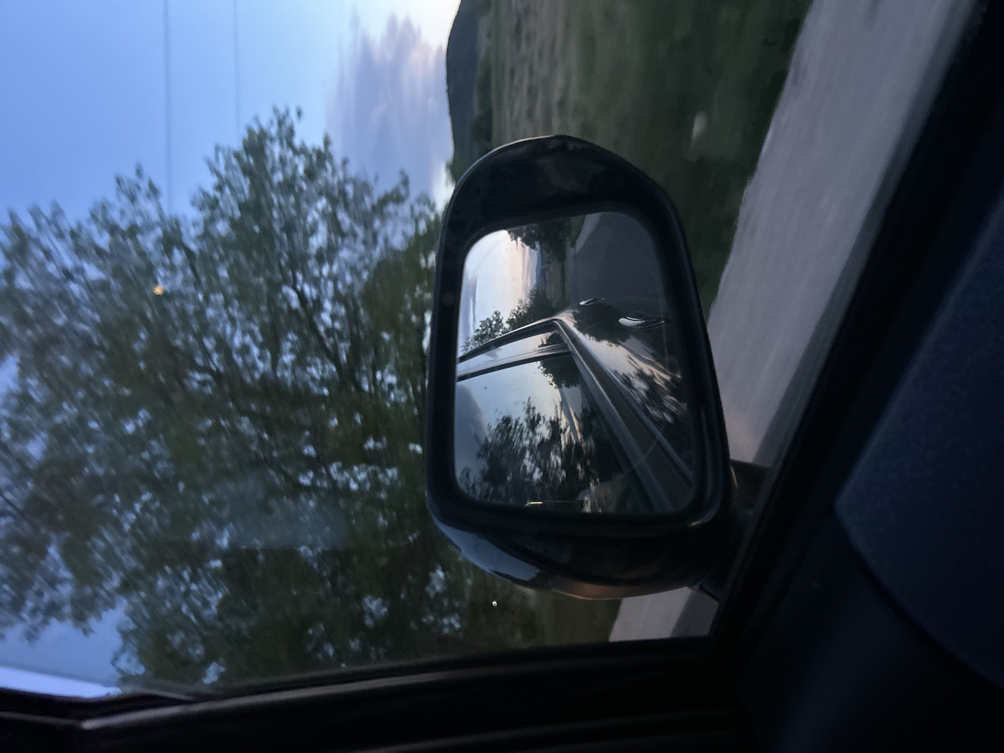 The image depicts a view from inside a moving vehicle, likely a car, with the side mirror prominently visible. The mirror reflects the road behind the vehicle, suggesting it's a highway or paved road. Outside the car window, there's a blurred view of trees and a hill or mountain in the distance. The sky is visible with some clouds. It is dusk or dawn, suggested by the lighting. The location is described as Ovchepoltsi, Bulgaria. No people are directly visible.
