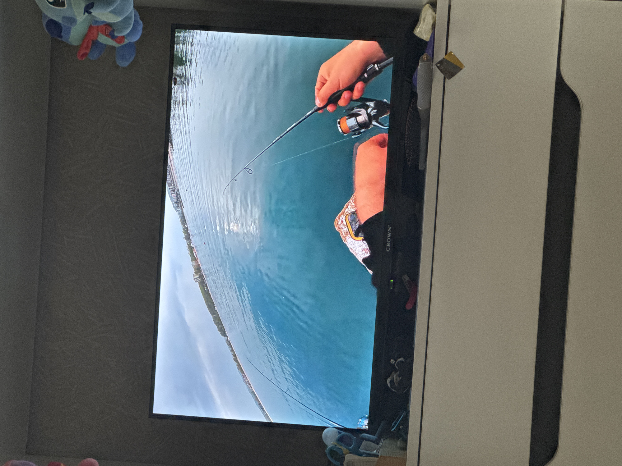 The image shows a TV screen displaying a video of a person fishing. The person is holding a fishing rod and reel over a body of water, likely a lake or sea, with a coastline visible in the background. The water is blue, and the sky appears overcast. The TV is placed near a wall, with a stuffed animal visible at the top left of the screen. The scene is indoors, and the surroundings include furniture, possibly a cabinet or closet. The location is impossible to be determined with only the information available.