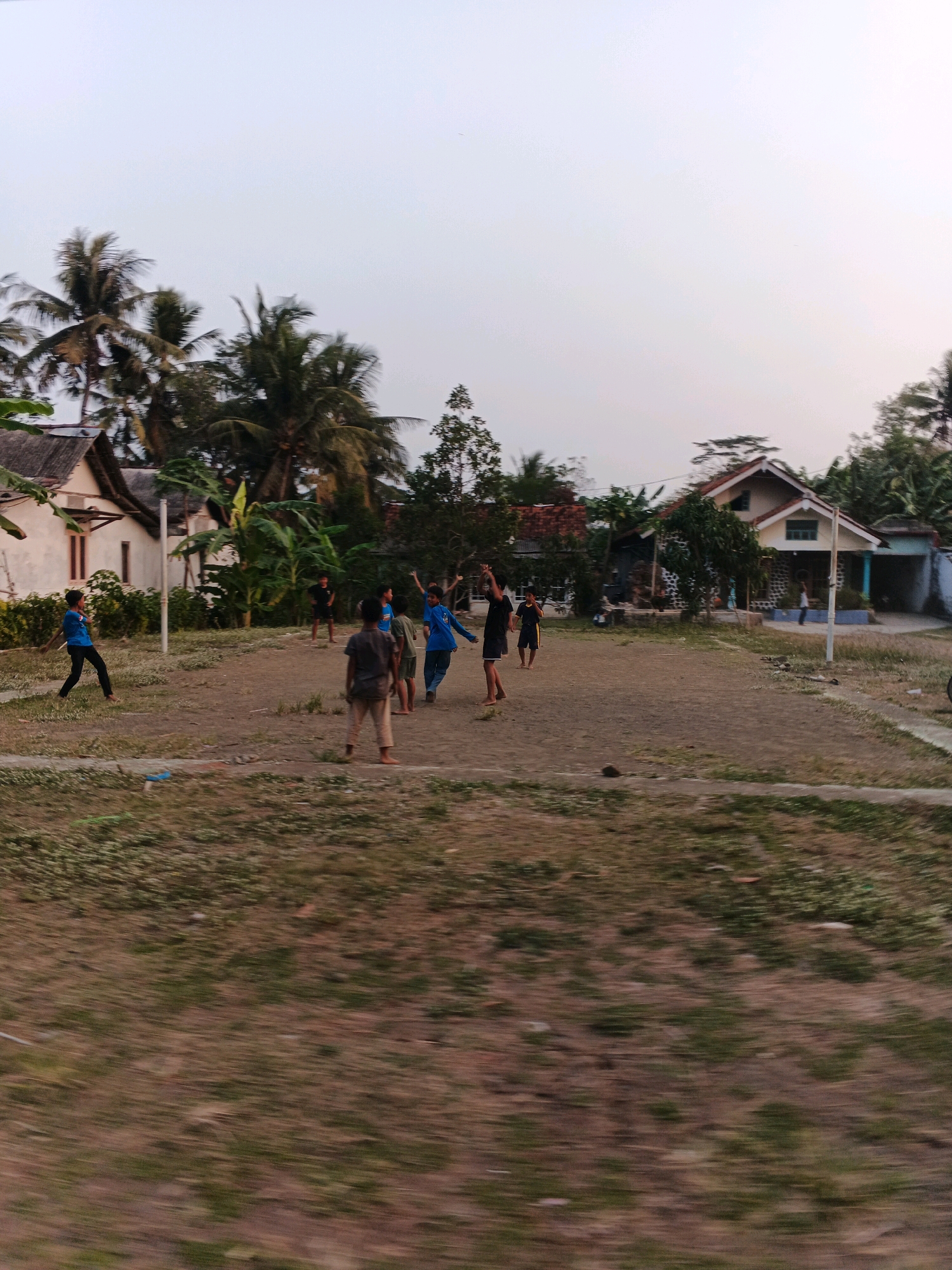 This image captures a lively scene in the village of Kesugihan, Indonesia, where a group of children are actively engaged in a game of volleyball on a dirt court. The children, varying in age, are dressed in casual clothing suitable for play. One child in a blue t-shirt and black pants is positioned on the left side of the court, in the process of kicking or hitting a ball. In the center, a boy in a gray shirt and light-colored shorts stands facing away from the camera, while another child in a blue shirt and shorts is in motion, possibly mid-jump or receiving a serve. Several other children are visible around the court, with some standing and others in active play.
The environment is characteristic of a rural Indonesian setting. To the left, a white house with a gray roof is partially visible, bordered by lush green vegetation and tall palm trees that dominate the background. Across the court, more houses, some with red tiled roofs, are nestled among dense trees and banana plants, suggesting a residential area. A volleyball net, indicated by poles and a visible net in the background, is set up on the court. The ground is a mix of packed dirt and patches of dry grass. The sky above is overcast, with a pale, hazy light suggesting it is either early morning or late afternoon. The overall impression is one of casual recreation and community life in a tropical village.