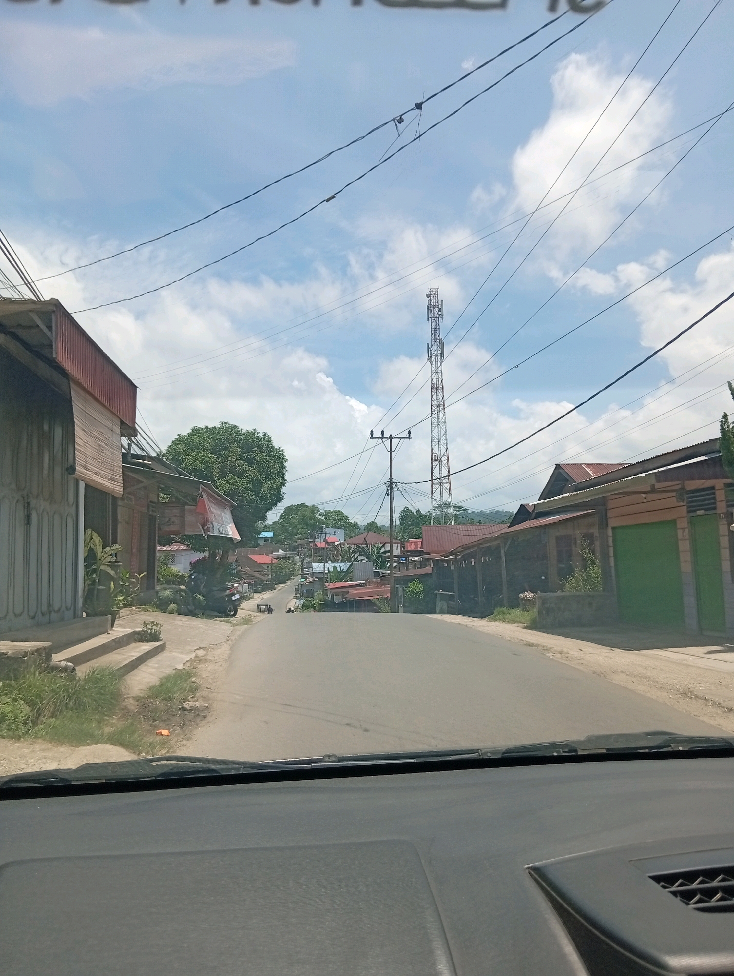 The image shows a road in the city of Gunungsitoli, Indonesia. The road is paved and appears to be a two-lane street lined with buildings on both sides. The buildings are a mix of residential and commercial properties, with some featuring corrugated metal roofs. A tall telecommunications tower is visible in the distance, along with power lines crisscrossing the sky. The sky is partly cloudy, indicating fair weather. There are a few motorcycles parked along the side of the road, and some vegetation, including trees and plants, is present. The photo appears to be taken from inside a vehicle, looking out through the windshield, with the dashboard visible in the foreground. There are no people clearly visible in the scene.