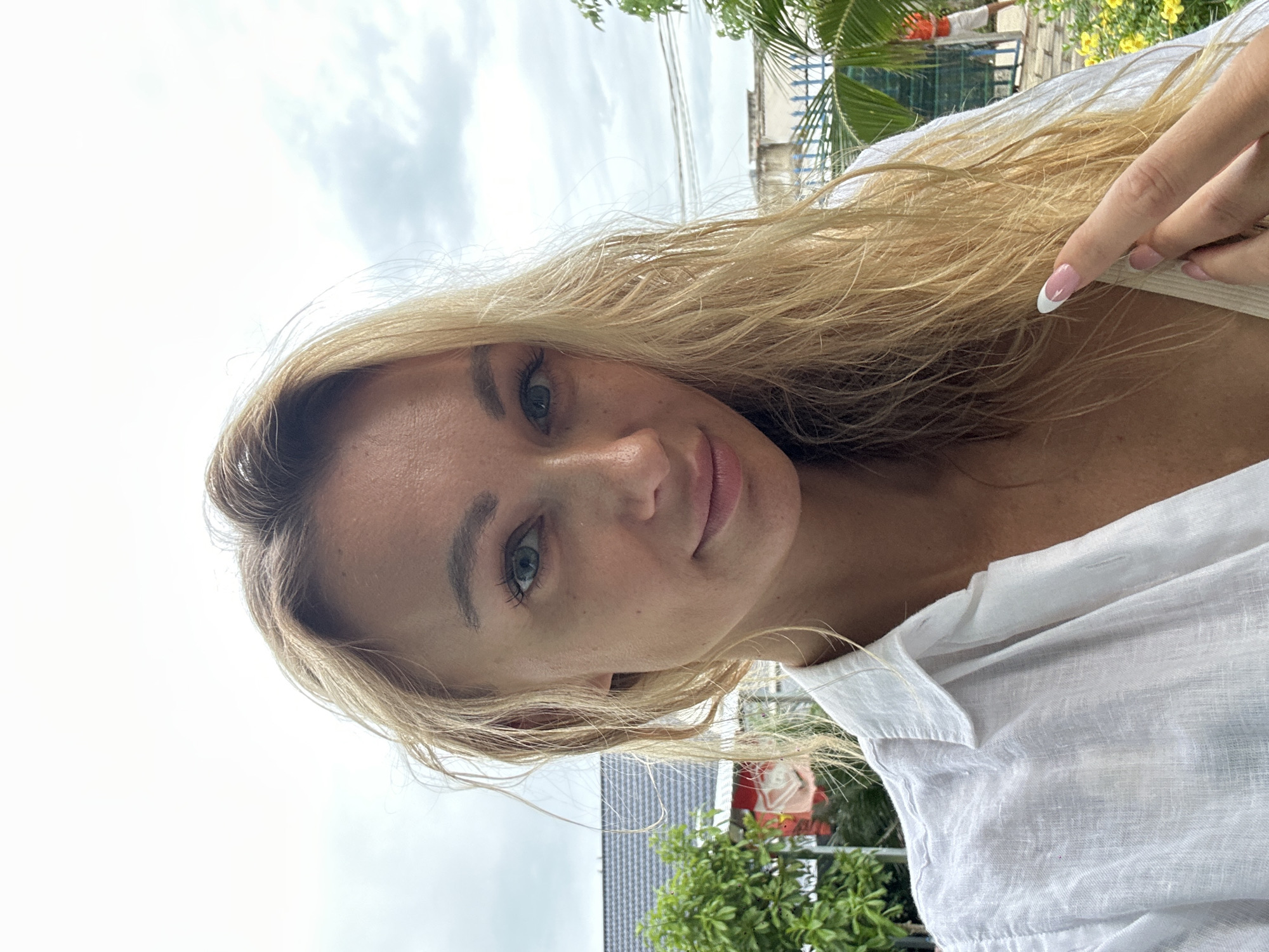 A close-up portrait of a woman with blonde hair wearing a white linen shirt. She is facing the camera with a slight smile. The background includes a cloudy sky, palm trees, and buildings. Based on the palm trees and architecture, the location is possibly in a tropical or subtropical climate, likely within Mũi Né, Vietnam. A finger with a French manicure is visible near her neck.