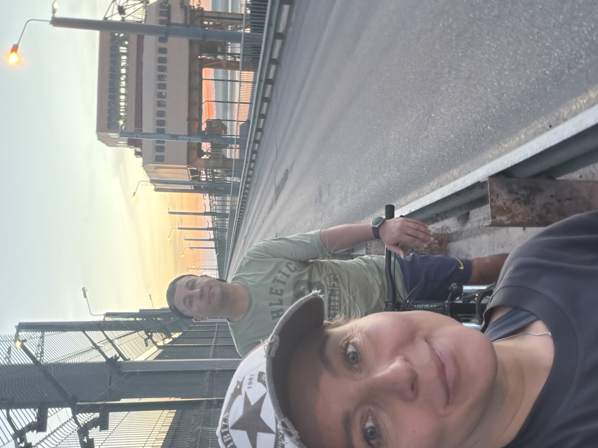 Two people are in the foreground, one closer to the camera, taking a selfie. They are on a bridge in Chaikovsky, Russia. A large building is visible on the other side of the bridge. The sky is light, suggesting it is early morning or late evening. The bridge has a metal barrier on the left side.
