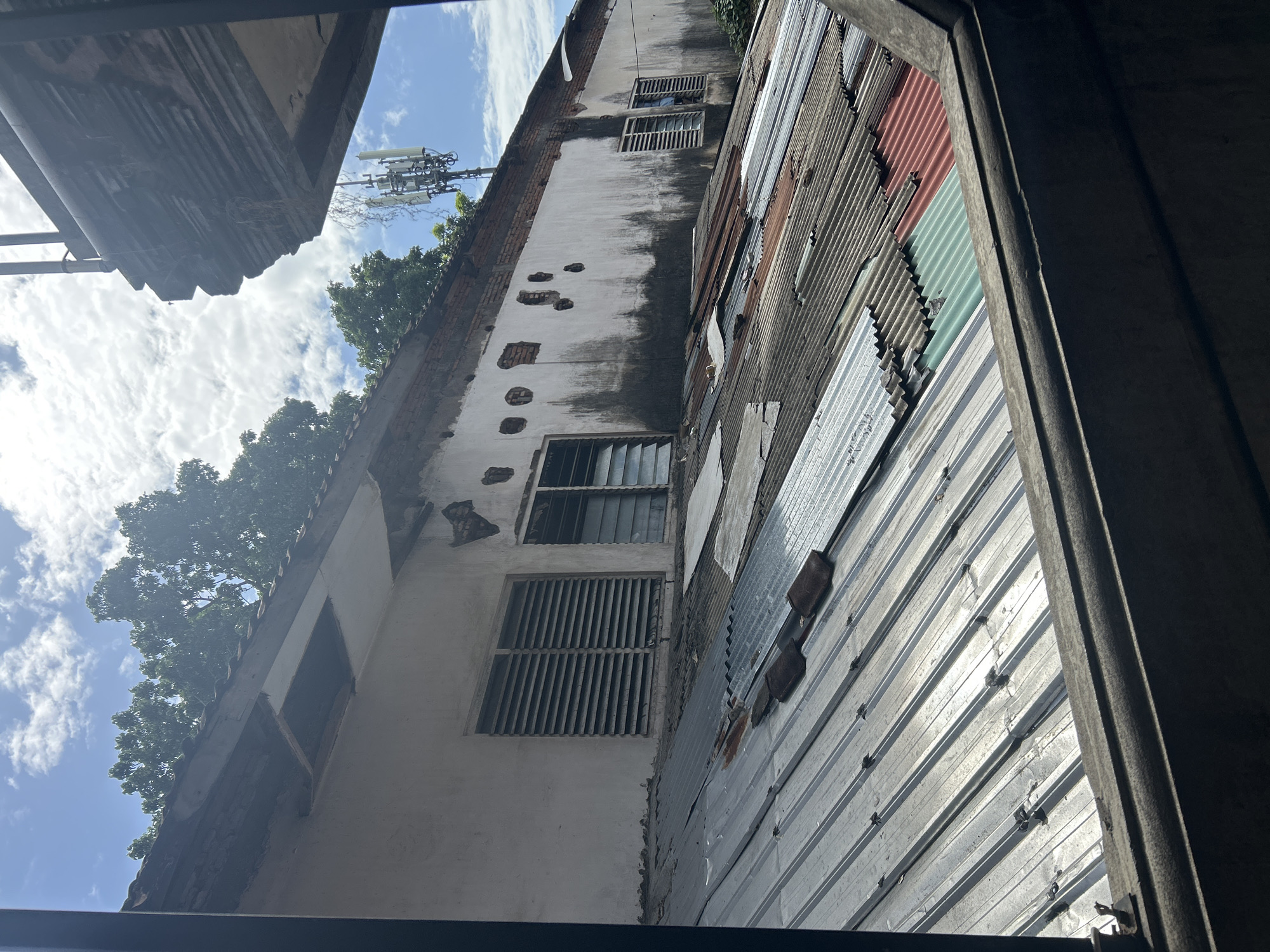 A partially obstructed view of a dilapidated building's exterior shows a section of a white plastered wall with exposed brickwork and dark staining, suggesting water damage or age. Two windows with horizontal louvers are visible on this wall. Below the wall, a corrugated metal roof slopes downwards. The roof is a patchwork of different materials and colors, including silver, rust-colored, green, and red corrugated metal sheets, some of which are damaged or missing. Several dark, rectangular objects, possibly roof tiles, are scattered on the roof. In the background, the sky is partly cloudy with a blue sky visible. A cell tower with antennas rises above the building's roofline. The scene appears to be in Denpasar, Indonesia, characterized by the humid climate implied by the building's condition and the tropical foliage visible in the upper left corner. The foreground shows a dark, textured ledge or frame, possibly from an adjacent structure or window.
