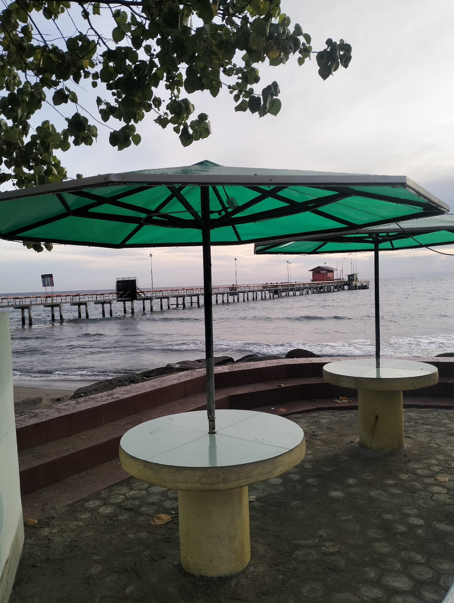 The image presents a tranquil coastal scene in Lhokseumawe, Indonesia, likely captured during the soft light of early morning or late afternoon under an overcast sky.
In the foreground, two round concrete tables, each with a light-colored top and a robust yellow-beige base, stand ready for visitors. Each table is shaded by a large, octagonal green umbrella, whose internal structure is visible. To the left, a portion of a light-colored wall or pillar is present, while a series of dark reddish-brown, tiered steps or seating areas recedes towards the water. The ground beneath these elements is a dark, coarse surface, possibly a mix of concrete and sand, with a few fallen leaves.
Beyond the immediate seating area, the calm ocean stretches out, with gentle waves breaking against a sandy and rocky shoreline. A long, sturdy pier or jetty extends prominently from the left side of the frame into the water, supported by numerous dark piles. Along the pier, several structures are visible, including a small dark building or platform closer to the shore, and a more distinct red-roofed building further out. Streetlights are interspersed along the length of the pier.
The sky is a soft, muted grey, indicating an overcast day. In the upper left corner, dark green leaves from an overhead tree partially frame the sky. No people are visible, lending a sense of peaceful solitude to the scene. Near the beginning of the pier, there is a sign with vertical columns of text, but the specific words are not discernible due to the distance and resolution. The overall atmosphere is one of quiet relaxation by the sea.