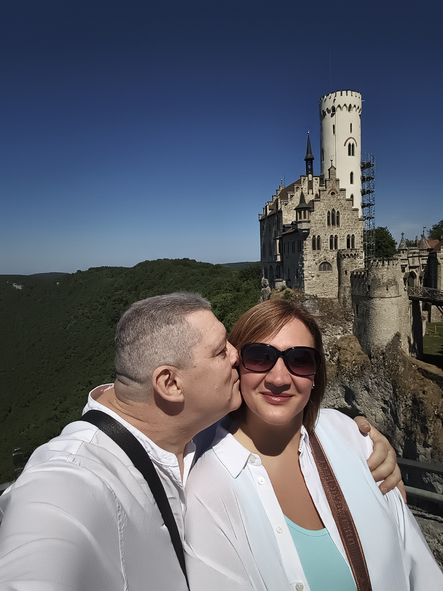 A couple is taking a selfie in front of Lichtenstein Castle in Honau, Germany. The man on the left is kissing the woman on the cheek. The woman is wearing sunglasses and a white shirt over a teal top. The man is wearing a white shirt. The castle, a prominent landmark, is situated on a cliff overlooking a lush green forest. The sky is clear and blue.