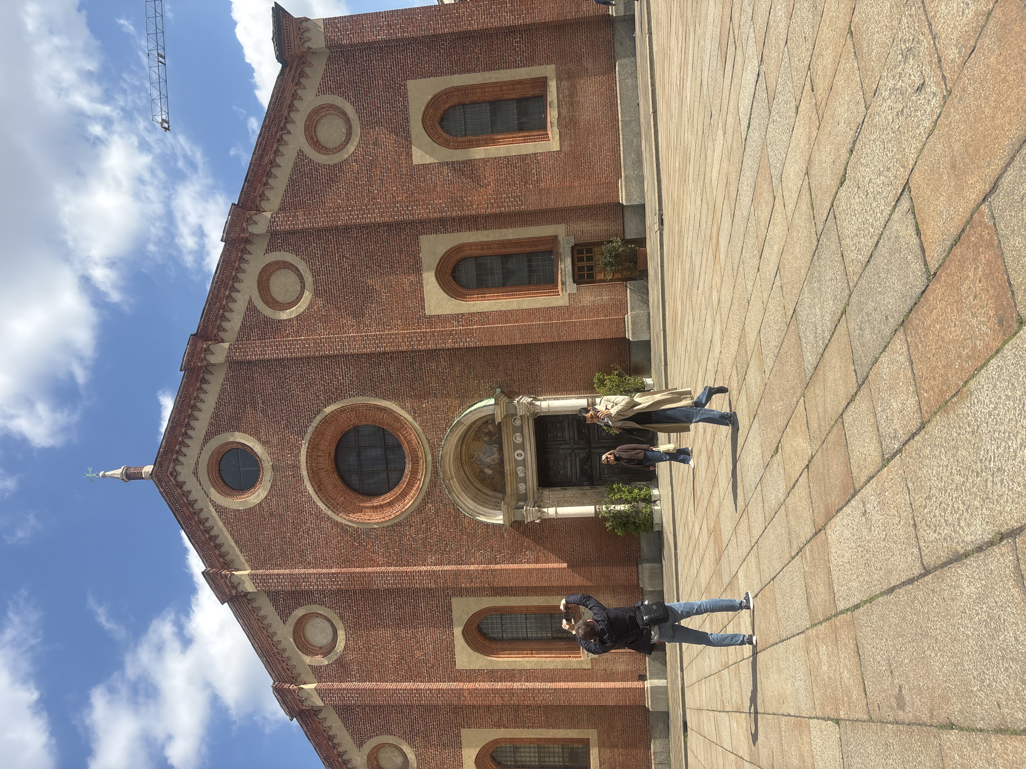 The image shows the Santa Maria delle Grazie church in Milan, Italy. The church is made of red brick with circular and arched windows. Three people stand in front of the church. One person is taking a picture of the church with a mobile phone, while the other two are standing near the entrance. The ground is paved with stone. A crane is visible above the church. The sky is blue with some white clouds.