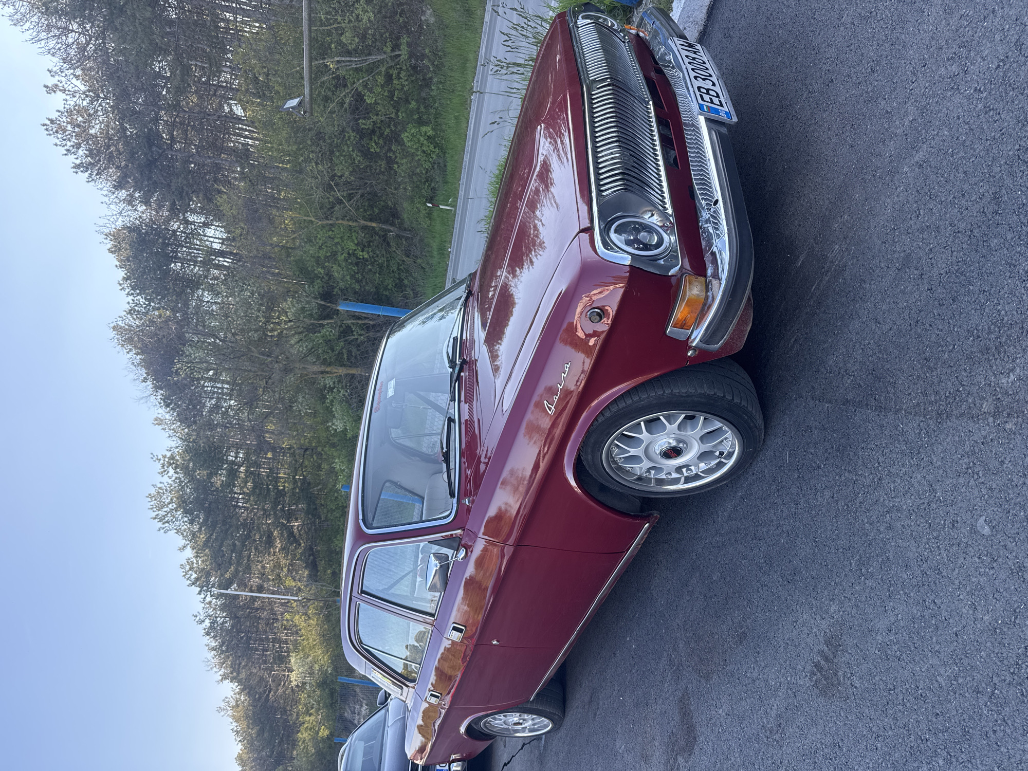 The image shows a maroon-colored GAZ-24 Volga sedan parked on an asphalt surface. The car has chrome detailing on the grille, headlights, bumpers, and window trim. The car's license plate reads "EB 3008 AM". The car is parked near a forest, under a blue sky. Considering the license plate registration and the type of vehicle, the location is likely in Varna, Bulgaria.