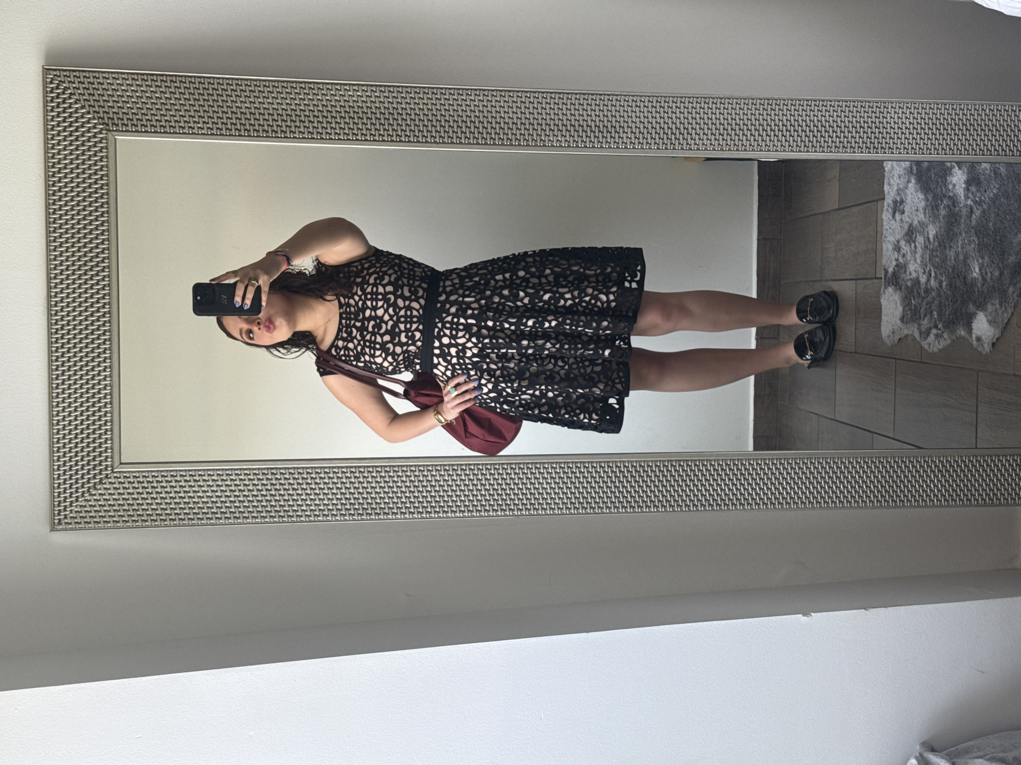 An adult female is captured in a full-length mirror selfie within an indoor setting. She stands with her left hand on her hip and holds a black smartphone in her right hand, obscuring her face from mid-torso upward, angled to photograph her reflection. Her lips are pursed. She wears a sleeveless black dress with a cut-out, patterned overlay revealing a light-colored slip or lining, cinched at the waist with a black band. Her attire includes black backless loafers with gold-toned hardware, and a burgundy shoulder bag rests on her left side. Visible jewelry includes blue nail polish, a gold-toned bracelet on her right wrist, and multiple rings, notably one with a light blue or green stone on her right middle finger. The environment consists of plain light-colored walls and dark brown rectangular floor tiles. A small, irregular gray shag rug is positioned on the floor in the foreground. The mirror features a silver-toned, textured frame with a woven pattern. A vertical wall protrusion is visible to the left of the mirror. The scene depicts a generic interior space, likely a residence, offering no specific architectural or environmental details to precisely pinpoint a location within Mexico City, Mexico.