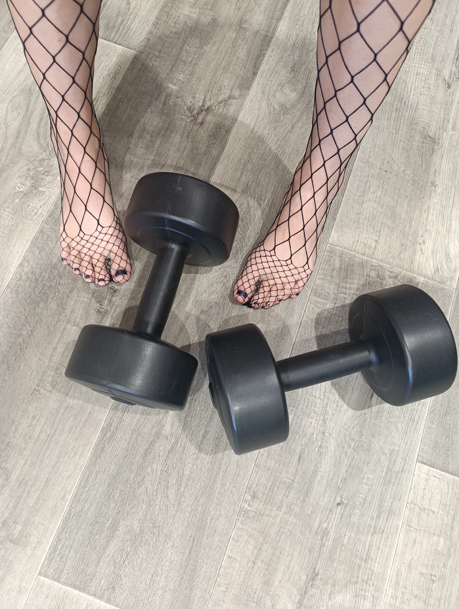 Two black dumbbells are on a light gray wood-grain floor. A person's feet, wearing black fishnet stockings and dark nail polish, are positioned above and between the dumbbells. The scene does not provide enough information to determine a specific location in Novokuybyshevsk, Russia.