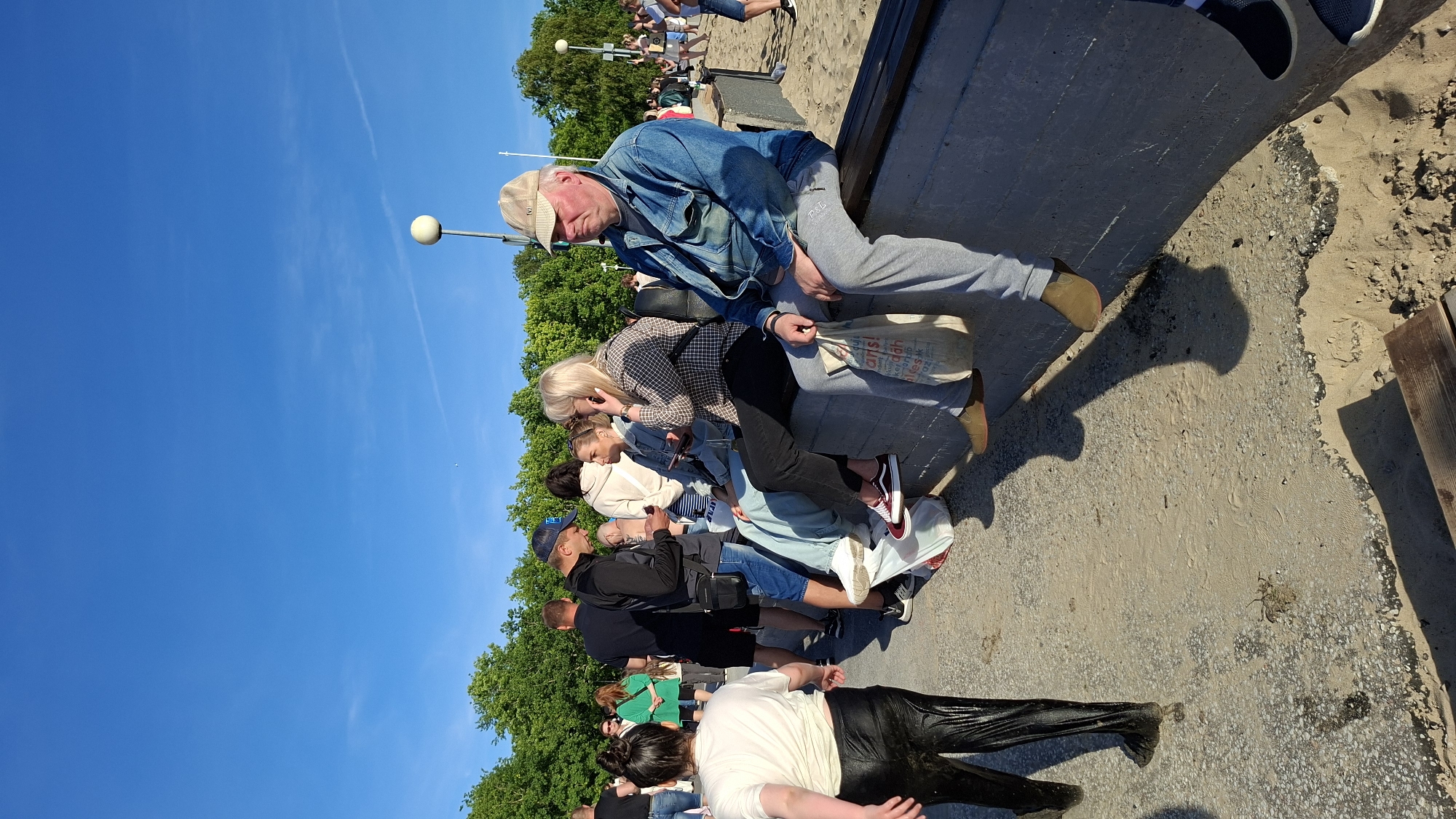 A group of people are gathered on a concrete ledge overlooking a sandy area. In the foreground, a person in wet, black pants and a white t-shirt walks away from the camera, their back to the viewer. To their right, several people are seated on the ledge.
One man, wearing a blue denim jacket and gray sweatpants with a cap, is seated in the center right of the frame, holding a white reusable shopping bag. To his left, a woman with blonde hair is on her phone, wearing a checkered shirt and black pants. Next to her, another woman in a light blue hoodie and jeans is looking at her phone. Further to the left, a man in a dark jacket and blue jeans is looking down at his phone and talking to someone. Behind him, other people are visible in the background, some standing and some seated.
The environment suggests a public outdoor space, likely a park or a waterfront area, given the sandy ground and the presence of trees in the background. The sky is a clear, bright blue. The scene appears to be taking place during daylight hours. The overall atmosphere is casual, with people engaged in conversation or simply relaxing. The location is likely near the coast in Tallinn, Estonia, possibly along the promenade or a recreational beach area.