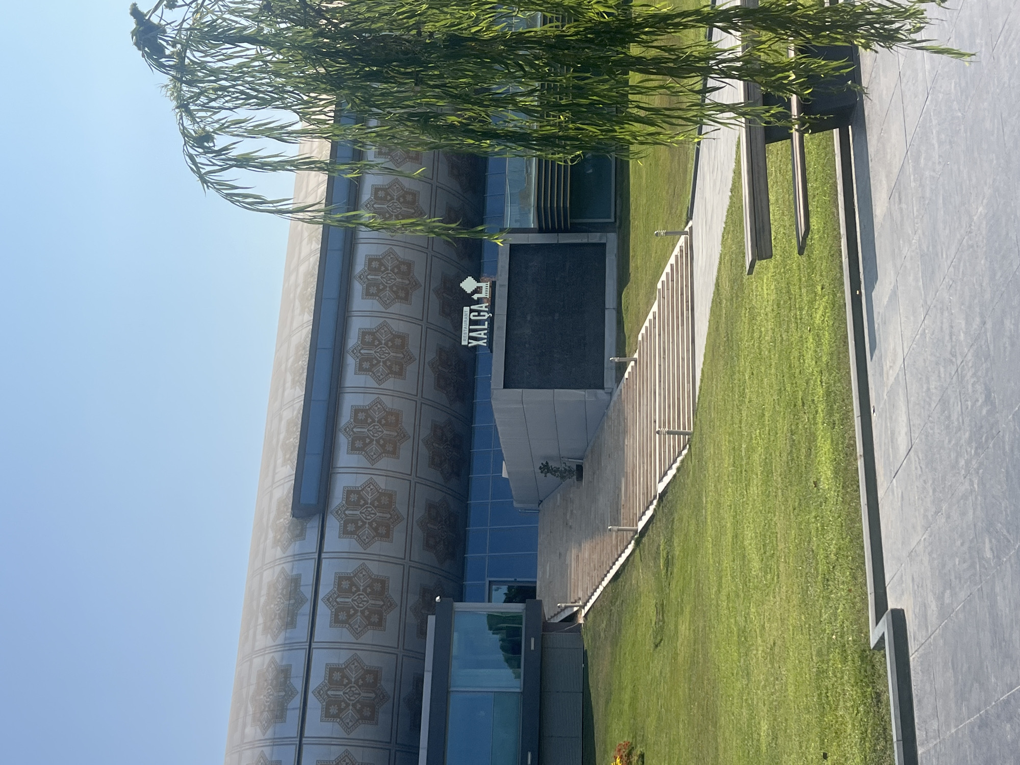 The image captures the exterior of the Azerbaijan Carpet Museum (also known as "Xalça" or "Xalca Museum"), a distinctive building located in Baku, Azerbaijan, on the embankment of the Caspian Sea. The museum's facade is adorned with repeating star-like patterns, and a prominent sign reads "RESTAURANT XALÇA". In the foreground, a lush green lawn slopes upwards towards a grand staircase leading to the museum's entrance. To the right of the staircase, a dark, rectangular panel, possibly a digital screen or a decorative wall, is visible. A weeping willow tree with long, pendulous branches partially obscures the right side of the building and the scene. In the lower right of the image, a modern-looking bench made of wood and dark material is situated on a paved area. The sky is clear and blue, indicating a sunny day. There are no people visible in the frame.