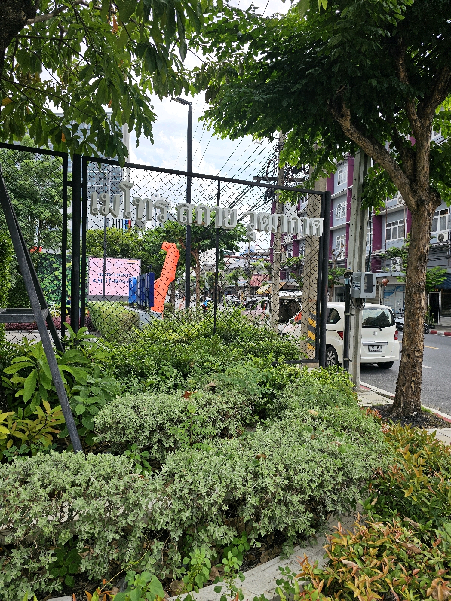 The image depicts a street scene in Bangkok, Thailand, with lush greenery in the foreground and a chain-link fence obscuring a view of a building and its surroundings. The fence is adorned with Thai lettering, which appears to be a name or signage. Behind the fence, a pink digital billboard displays the words "INSTALLMENTS 4,500 THB/mo." The billboard is flanked by abstract orange and blue sculptural elements. Beyond that, a white van is parked, and a tree with a thick trunk grows on the right side of the frame. The background shows a multi-story building with purple and gray facades and multiple windows. The sky is overcast.