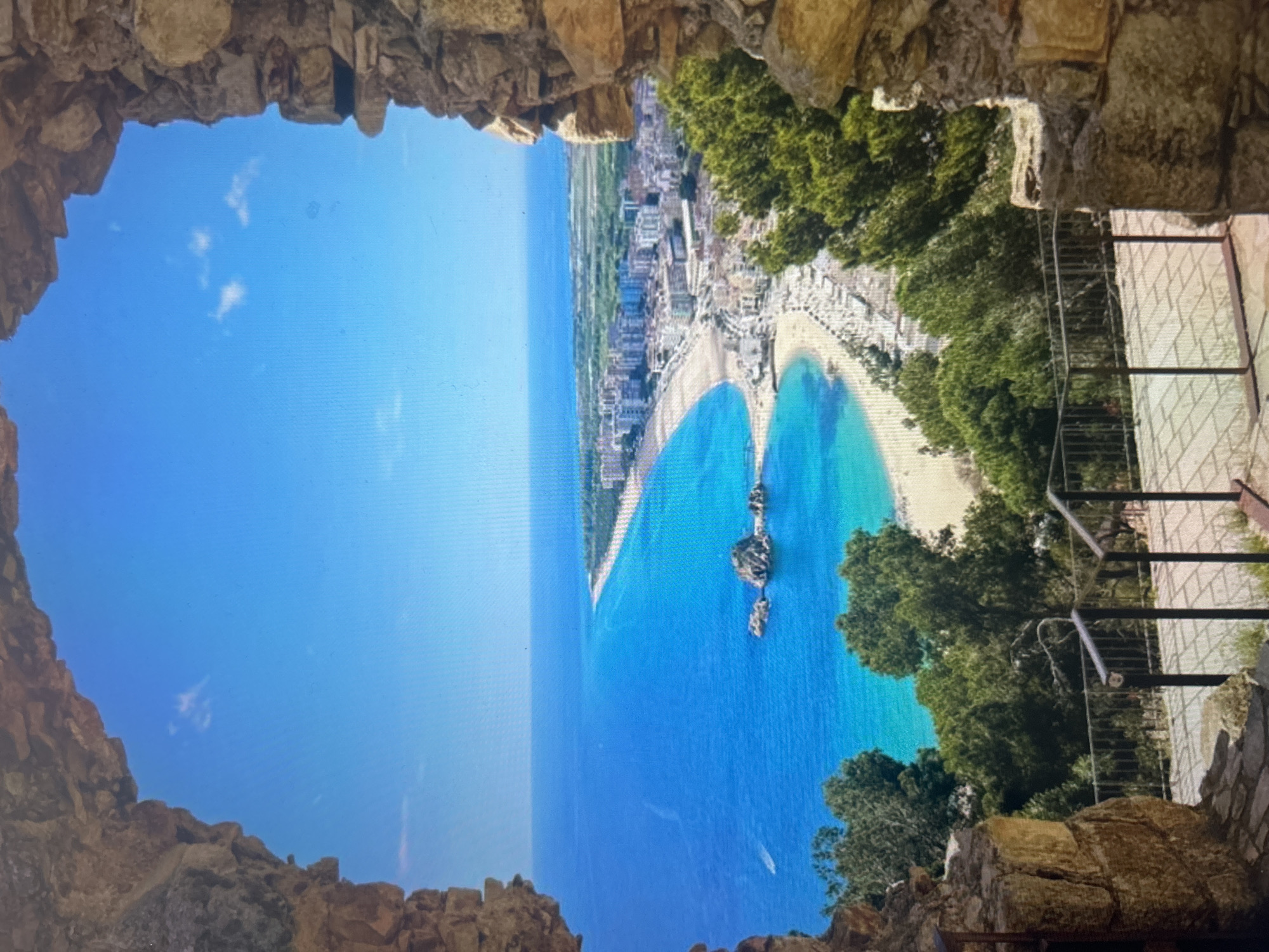 The image presents a breathtaking panoramic view of a coastal landscape, framed by a rugged, natural stone archway or opening in the foreground. The scene is brightly lit, suggesting a clear, sunny day.
Through the earthy-toned stone frame, a vast expanse of brilliant blue sky stretches across the top, adorned with a few delicate, wispy white clouds. Below the sky lies the deep blue ocean, transitioning to a vivid turquoise closer to the shoreline. The coastline features two distinct, crescent-shaped sandy beaches, gracefully curving to form a heart-like shape, separated by a prominent rocky outcrop or small island emerging from the clear waters. Another smaller rocky formation is visible further out in the bay.
Along the curved shoreline, a dense cluster of buildings forms a vibrant coastal town, extending into the distance. Lush green trees and foliage are interspersed throughout the landscape, particularly on the hillsides and near the edge of the stone frame on the right. On the far right, partially visible behind the rockwork, is a metal staircase with railings, suggesting a path or an elevated viewing platform. A tiny white boat can be faintly seen in the distant ocean.
The image captures a serene yet grand vista, likely from a high vantage point such as a castle or cliffside lookout, offering an idyllic view of a popular resort or vacation spot. There is no visible text in the image.