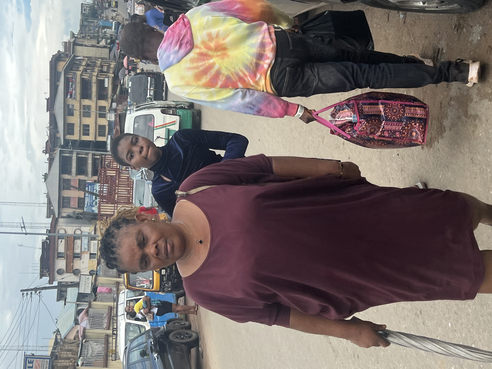 A medium shot captures three people walking on a street in Nkpor, Nigeria. In the foreground on the left, a woman with braided hair faces forward, wearing a burgundy dress and a silver necklace. She holds a coiled cable in her right hand. To her right, a young person in a dark blue top looks towards the camera. Further to the right and walking away from the camera is a person wearing a tie-dye hoodie in shades of yellow, pink, and blue, black jeans, and carrying a large patterned bag in their left hand and a black plastic bag in their right. In the background, various buildings with multiple stories line the street, with parked vehicles and a cloudy sky overhead. The scene appears to be a busy urban environment.