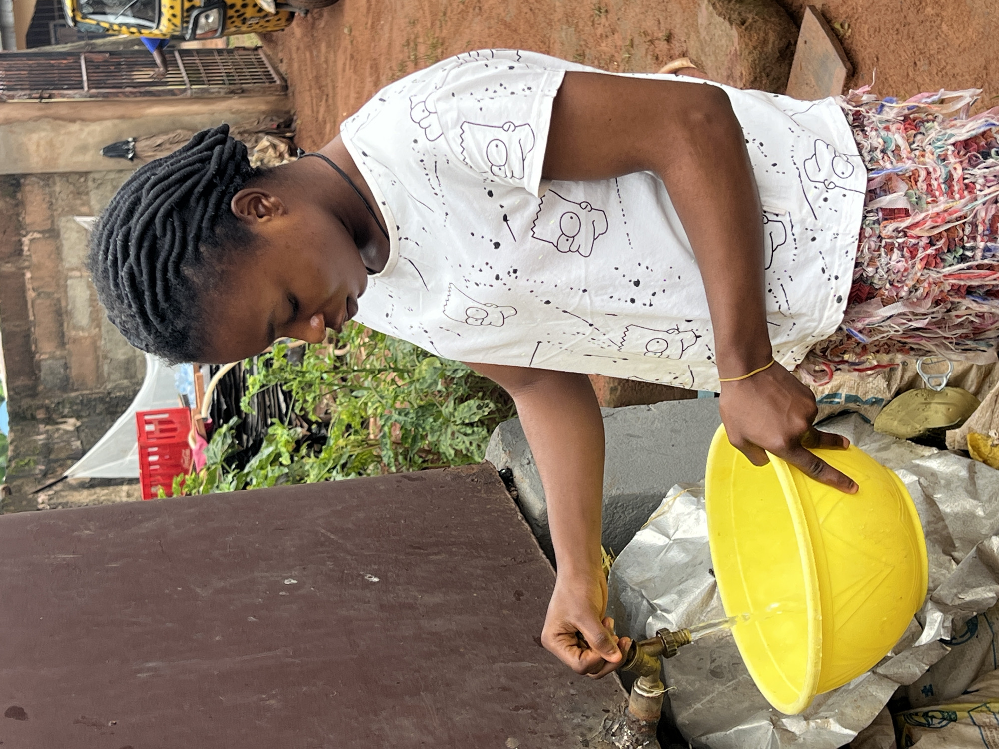 In Nkpor, Nigeria, a young woman with dreadlocks is collecting water from a faucet into a bright yellow basin. She wears a white t-shirt with a pattern of cartoon characters and a colorful, fringed skirt. The faucet is attached to a large, dark brown cylindrical tank, and the ground is dirt. Behind her, a yellow and black auto-rickshaw is visible through a metal gate, and stacks of red crates and greenery are present.