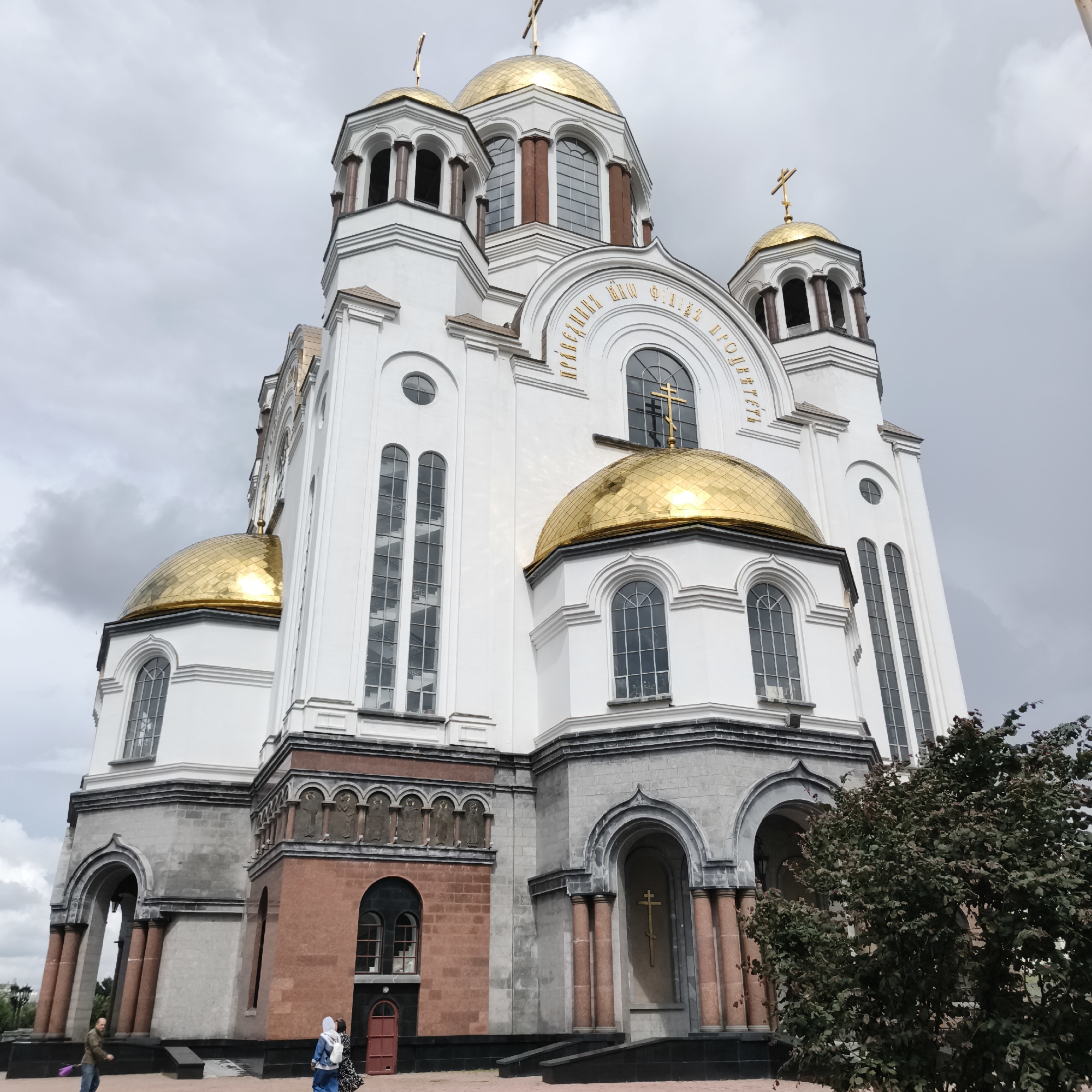 The image shows the Church on Blood in Yekaterinburg, Russia. The church is a white Orthodox cathedral with gold domes and crosses. The building features large arched windows and decorative detailing. The sky is overcast with grey clouds. In the foreground, a few people are visible. On the left, a man in a brown jacket and jeans is walking away from the camera, holding a purple object. On the right, two women are standing and looking at the church. One woman is wearing a blue headscarf and a blue jacket, carrying a white backpack. The other woman is wearing a patterned dress. There is also some greenery in the lower right corner of the image, appearing to be a tree or bush.