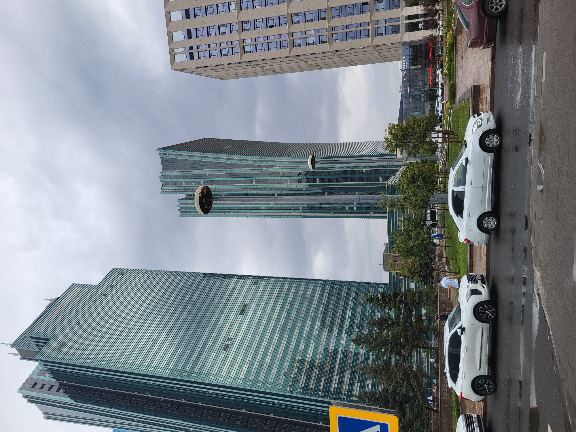 The image shows a street scene in Astana, Kazakhstan, featuring modern architecture and vehicles. In the foreground, a wet asphalt road is visible with puddles, indicating recent rain. Three cars are parked or driving on the road: a white SUV on the left, a white sedan in the center, and a dark red sedan on the right. A yellow and blue traffic sign is partially visible on the left side of the frame.
The background is dominated by several tall, contemporary buildings. To the left, a large skyscraper with a green-tinted glass facade rises high into the overcast sky. In the center, another distinctive skyscraper has a warped, green-and-grey patterned glass exterior. To the right, a rectangular building with a concrete-like facade and numerous windows is visible.
Between the buildings, there are trees and manicured green spaces, with pathways and more buildings in the distance, some of which appear to be commercial establishments with outdoor seating areas marked by red umbrellas. Several pedestrians are scattered throughout the scene, walking on sidewalks or near the buildings. Streetlights are also present, one of which is prominently featured in the middle ground. The sky is heavily overcast, suggesting a cloudy day.