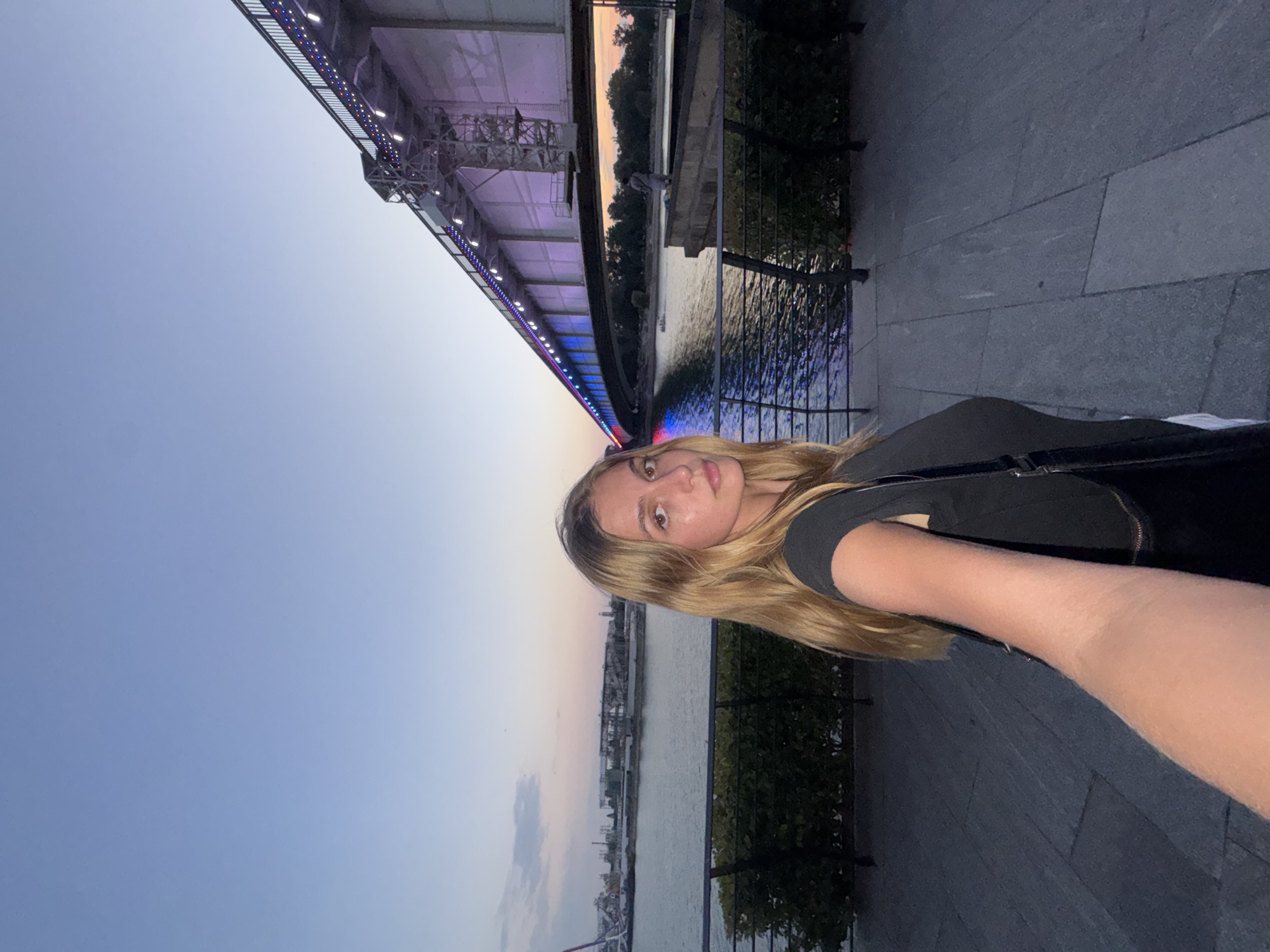 The image depicts a young woman with long blonde hair taking a selfie in an urban waterfront setting at dusk. She is wearing a dark t-shirt and carrying a black bag. Behind her, a large bridge is illuminated with colorful lights, casting reflections on the water. The bridge appears to be part of Belgrade's urban infrastructure, possibly the Gazela Bridge, given its distinctive design and lighting. The riverbank in the foreground features a paved walkway, low green hedges, and a railing. In the background, across the river, there are industrial structures and trees silhouetted against the twilight sky. The overall atmosphere is serene and captures a moment of leisure by the Sava River.