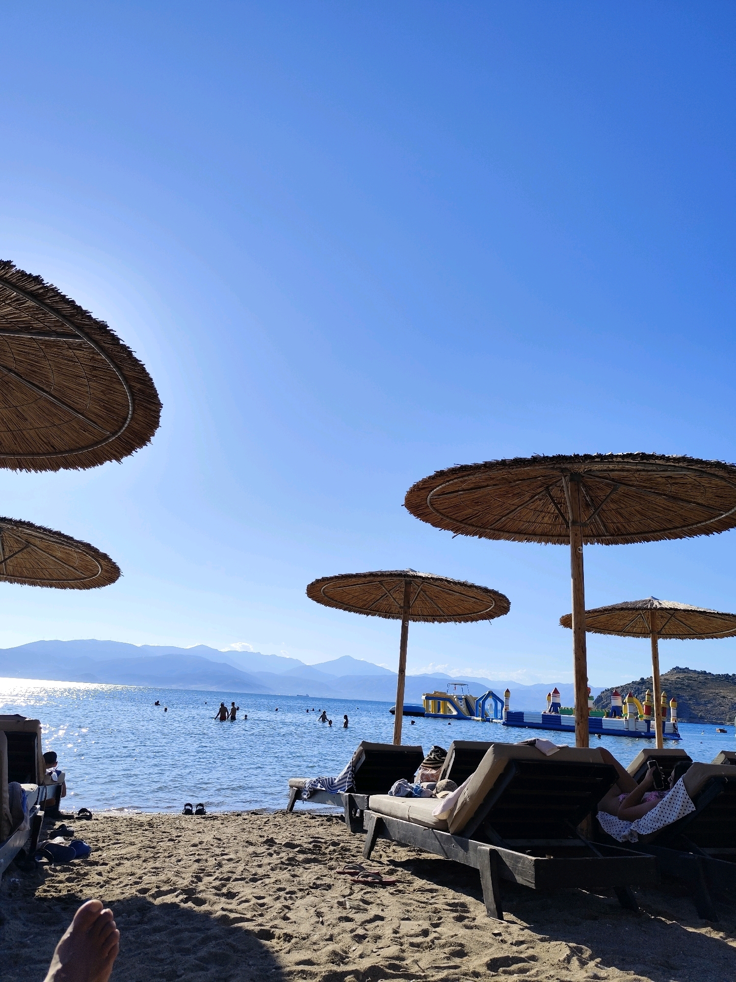 The image depicts a beach scene in the Municipal Unit of Nafplio, Greece, under a clear blue sky. In the foreground, sandy beach stretches out, with several empty lounge chairs casting shadows. One lounge chair in the mid-ground has a person reclining on it, looking at their phone. The sea is calm and blue, with several people swimming and wading in the water. In the distance, there is an inflatable water park on the sea, and mountains rise beyond the coastline. Several thatched umbrellas provide shade on the beach. The overall atmosphere is one of a tranquil summer day at the seaside.