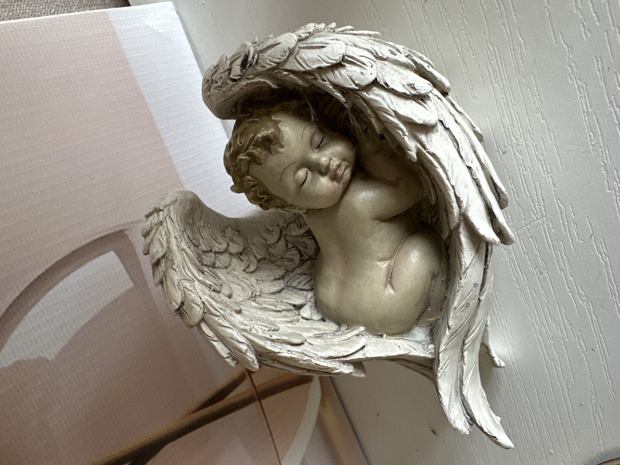 An angelic figurine of a sleeping cherub is placed on a white, textured surface. The figurine is light-colored, with detailed feathered wings curled around the cherub. The cherub's eyes are closed, and its head is resting on its arm. In the background, a pale pink and white textured surface is visible, with a hint of a shadow. The image is a close-up, focusing on the figurine and its immediate surroundings. There are no people or specific landmarks of Rogozkino, Russia, identifiable in this image.