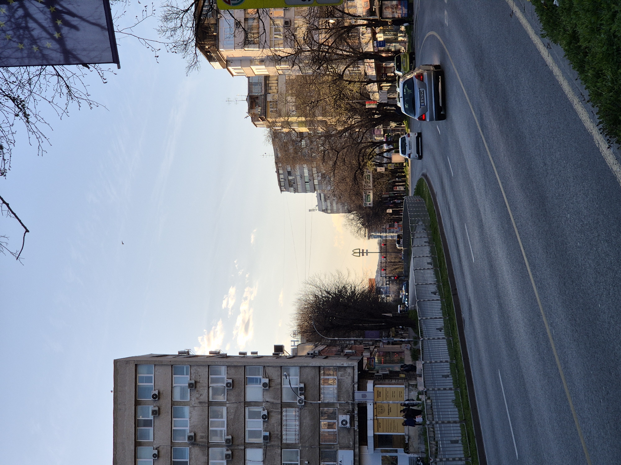 The image depicts a street scene in Varna, Bulgaria. A wide road with two lanes runs diagonally across the frame. On the right side of the road, there is a green curb and buildings on the other side of the road. Silver car is visible in the road. On the left side of the road, there is a multi-story concrete building with multiple windows and air conditioning units. Buildings run along the horizon of the image. A European Union flag is visible in the upper left corner. The sky is clear and light blue.