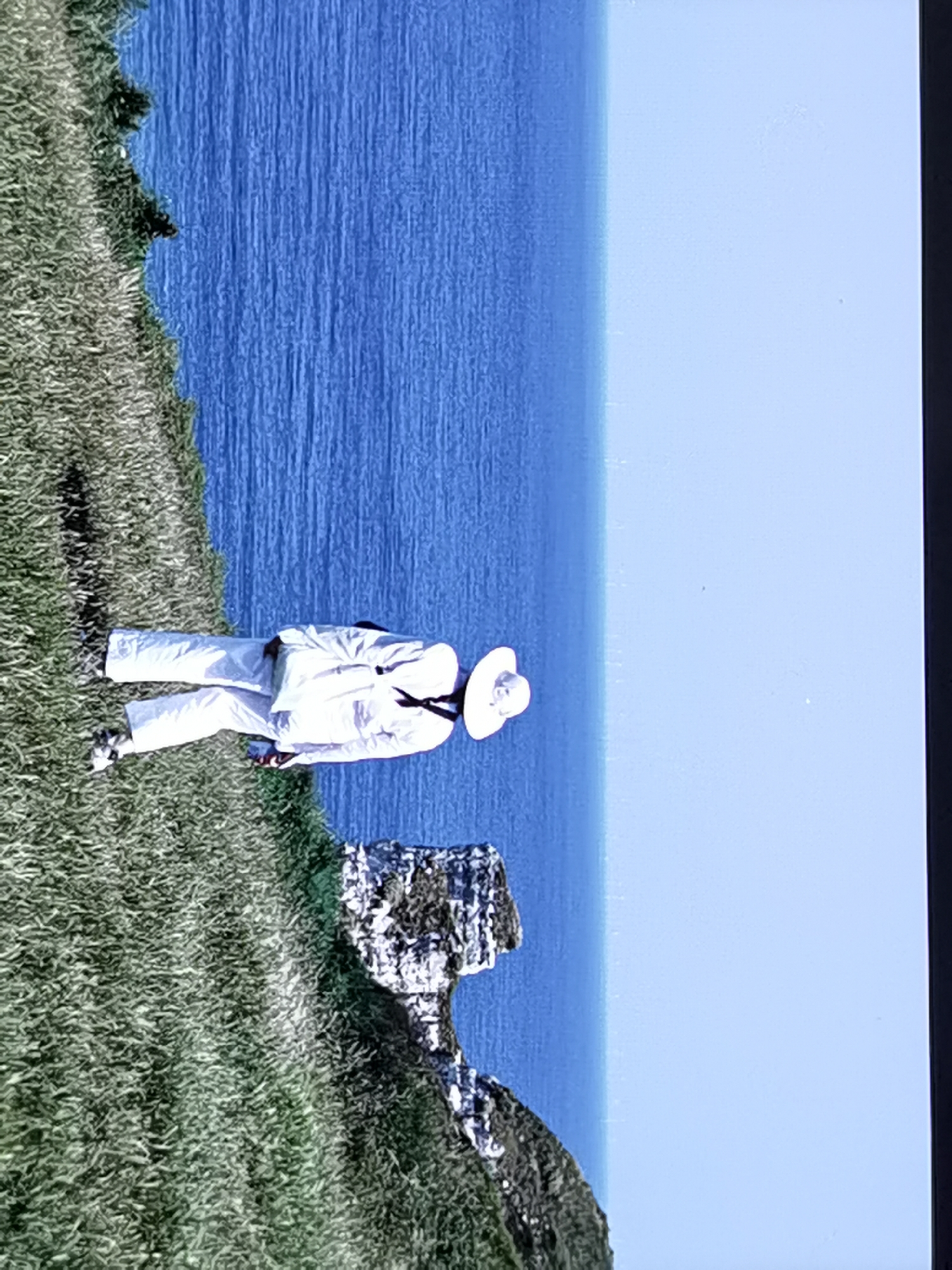 The image shows a person standing on a grassy cliff overlooking a blue ocean. The person is wearing a white suit, a white hat, and dark shoes. The person's back is to the camera, and they are looking out at the ocean. The scene appears to be set during the day, with clear skies and calm water. The grassy cliff suggests a coastal location, and the rocky outcrop in the foreground is a characteristic feature of the cliffs at Étretat, France. The overall mood of the image is peaceful and contemplative, with the vastness of the ocean and the natural beauty of the landscape creating a sense of serenity. There is no visible text in the image.