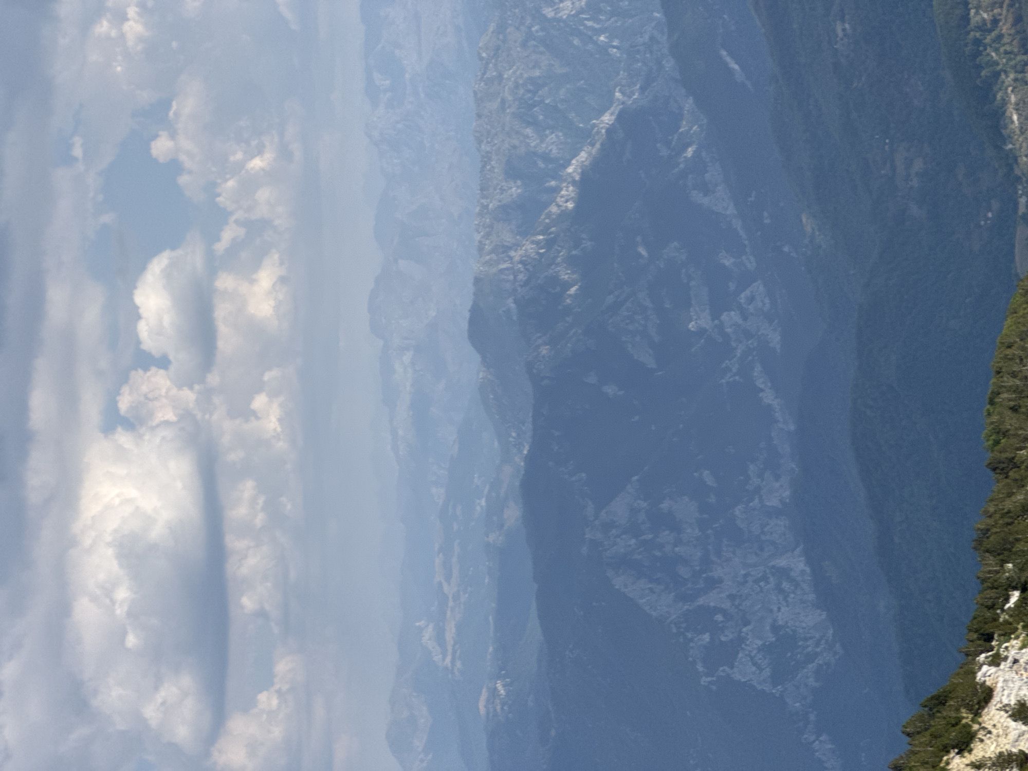 The image depicts a vast, layered mountain landscape under a sky filled with large, fluffy white and grey clouds. The mountains recede into the distance, becoming progressively hazier and bluer with depth. The foreground shows a steep, rocky slope with scattered green trees. There are no people or discernible man-made structures visible in the main body of the image. The scene is a natural, mountainous environment, consistent with the topography of Mrtvo Duboko, Montenegro.