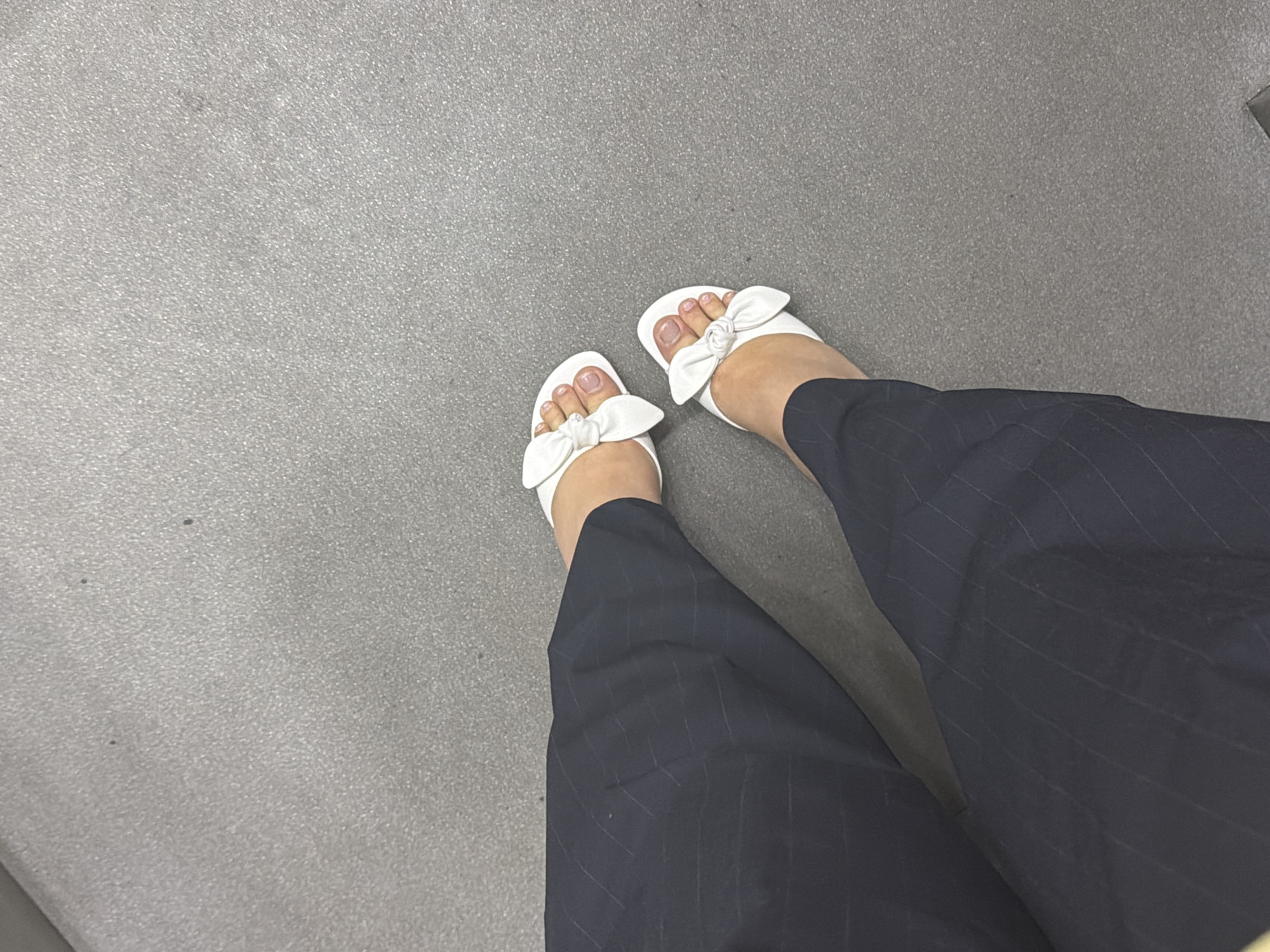 The image shows a first-person, aerial view of a person's lower extremities. The person is wearing dark-colored pinstripe pants and white sandals with bow embellishments. The sandals expose the person's toes, which are adorned with a light pink nail polish. The person is standing on a gray, smooth surface. The location appears to be indoors, possibly a room with a plain, utilitarian floor. There are no indications within the image to associate it with Yerevan, Armenia.