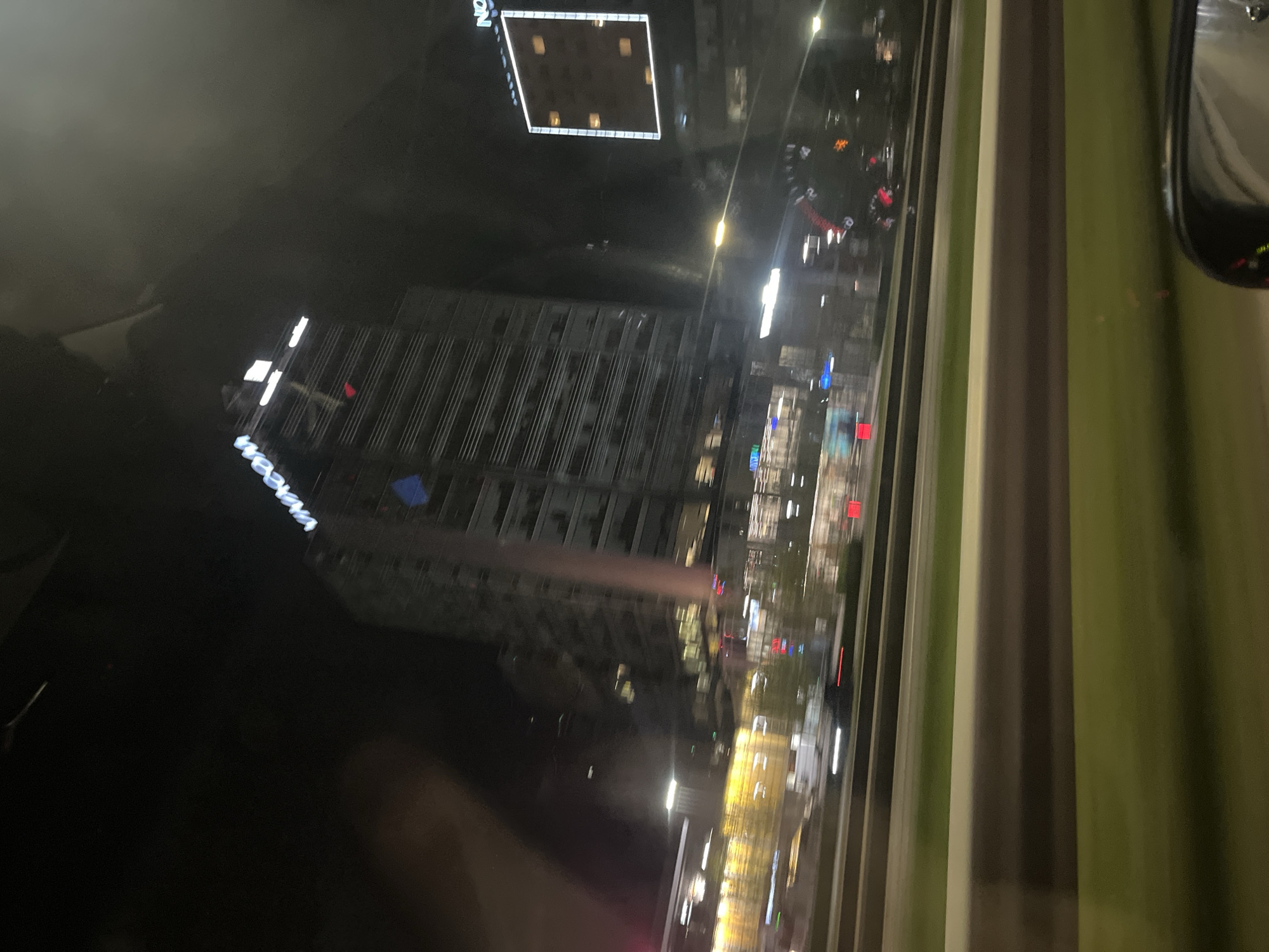 The image, rotated approximately 90 degrees clockwise, captures a nighttime urban scene from the interior of a moving vehicle, likely in Sofia, Bulgaria. Significant motion blur across the frame underscores the vehicle's movement.
On the left, reflections on the window obscure parts of the view, showing indistinct shapes from inside the vehicle. A prominent multi-story building, characterized by vertical architectural lines, dominates the mid-ground. On this building, a white illuminated sign displays partially discernible letters, appearing to start with "wes" or similar, distorted by reflections and the angle.
Further in the background, another tall structure features a large, brightly illuminated square sign. Within its top-left corner, the letters "ON" or "NO" are visible. Numerous other artificial lights from streetlamps, other buildings, and possibly other vehicles dot the dark landscape, appearing as luminous streaks due to the motion.
The right side of the image reveals elements consistent with a road or adjacent infrastructure, including vertical green and grey stripes that could be road barriers, a median, or even curtains within the vehicle. A portion of a vehicle's side mirror is clearly visible in the upper right corner, reinforcing the perspective from inside a car, bus, or similar transport. The overall scene conveys the experience of traveling through a city at night.