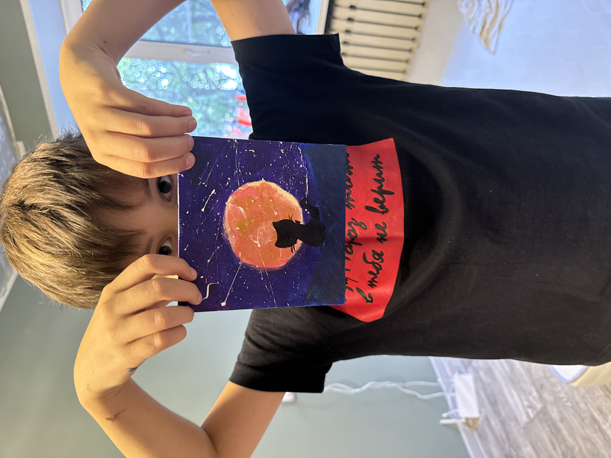 A young person with brown hair is holding up a painting in front of their face, obscuring most of it. The painting depicts a starry night sky with a large orange moon and a silhouette of a black cat sitting on a hill. Below the painting, a red band across the person's black t-shirt has handwritten text in black. The background shows a room with light-colored walls and wooden flooring, and a window with a view of trees and foliage. A white radiator is visible along the wall. The overall scene suggests an indoor setting, possibly a home or studio in Cheboksary, Russia.