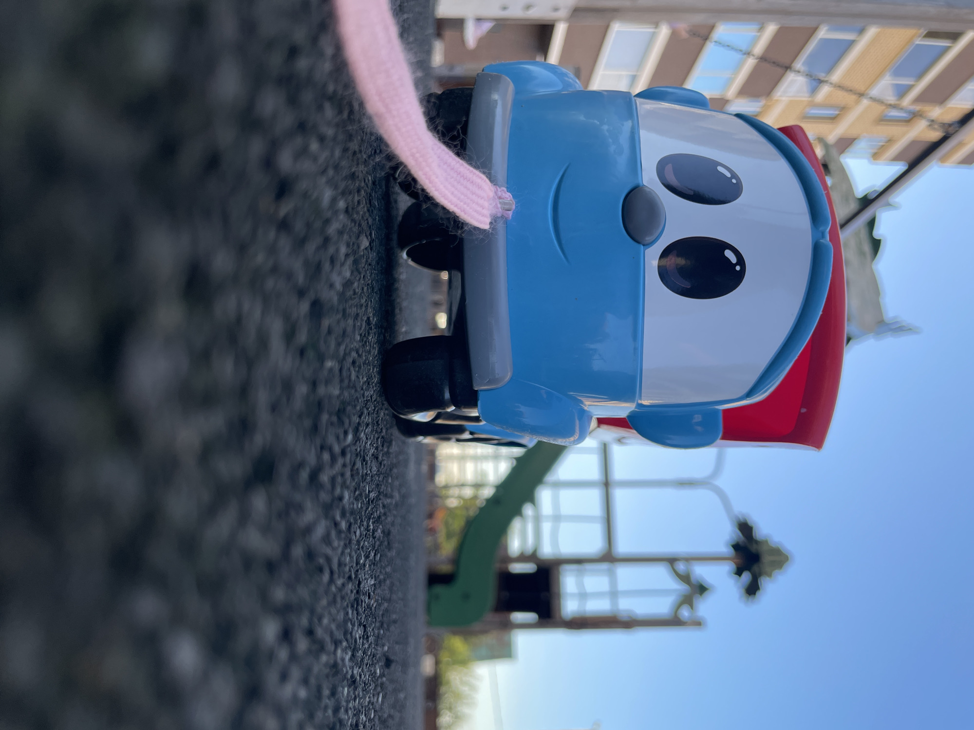 A low-angle photograph taken in Izhevsk, Russia, presents a blue and white toy truck with a red cargo bed positioned centrally on a dark, textured playground surface. The toy truck's front face displays two large black eyes with white reflections, a grey oval nose, and a simple black smile. A pink, braided string or shoelace is securely tied to the center of the toy truck's front bumper, extending forward and slightly out of frame towards the viewer. In the blurred background to the left, a multi-story residential building with a light brown facade and numerous windows is visible. To the right, a playground structure featuring a green slide and metal supports is indistinctly rendered. The upper portion of the frame shows a clear, bright blue sky. The scene is illuminated by natural daylight. No individuals are present in the image.