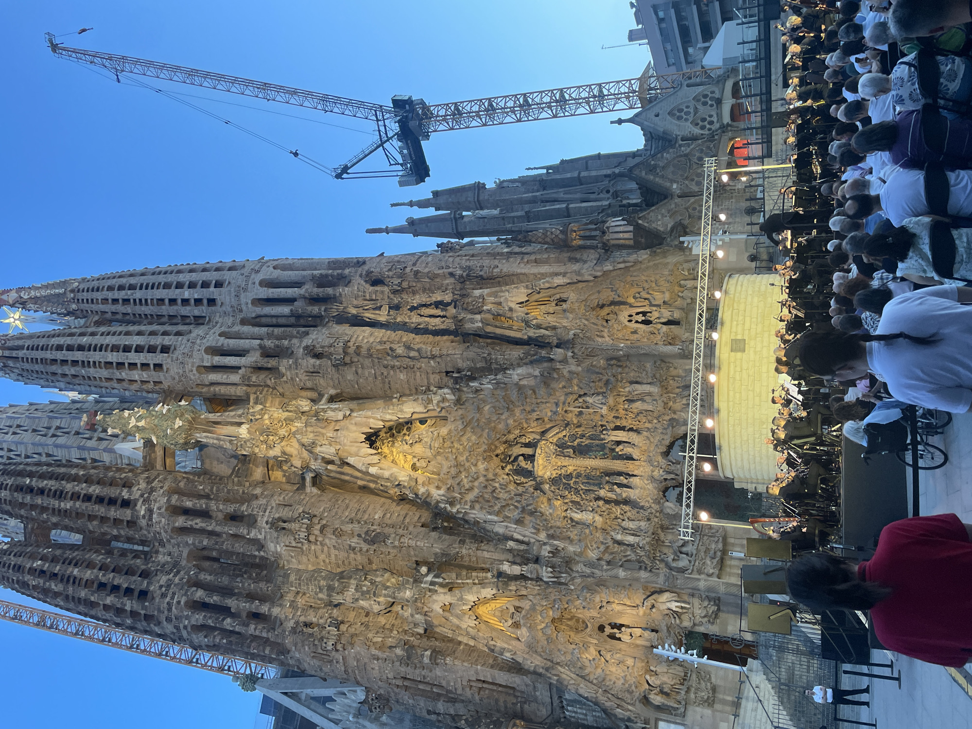 The image captures a twilight scene in front of the Sagrada Familia basilica in Barcelona, Spain. The basilica's facade, intricately carved and partially illuminated, dominates the background. A large construction crane looms over the right side of the structure, indicating ongoing work. In the foreground, an orchestra is performing on a stage set up outdoors. Numerous people are seated, attending the performance. Some individuals are standing, observing the scene. The sky is a clear, deep blue, transitioning into twilight. The overall atmosphere suggests an outdoor concert or event taking place in a prominent urban location.