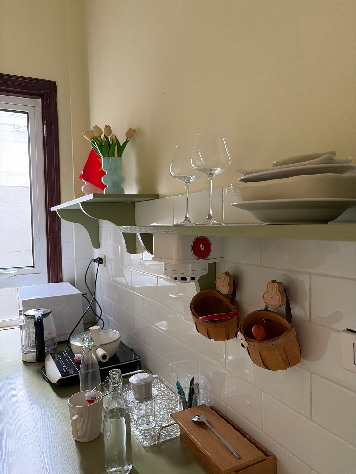 The image depicts a kitchen counter with white tiled walls and light green shelves. On the top shelf, there are two wine glasses and a stack of plates. On the bottom shelf, there is a white device with a red circular button, possibly a smoke detector or a smart home device. Below the shelves, on the counter, are various kitchen items. To the left, there is a white electric kettle and a black induction cooktop with a pot on it. Next to these are two glass bottles with stoppers, one filled with water. A beige mug and a small container are also present. Two woven baskets with bunny-shaped wooden handles hang on the wall, one containing a red object and the other a reddish-orange object. A wooden box with a spoon resting on it is also visible. The overall impression is of a neat and organized kitchen space. The presence of a Vietnamese flag in the background, partially visible on the top shelf, along with the distinct design of the coffee dripper on the counter, strongly suggests this scene is located in Hà Nội, Vietnam.
