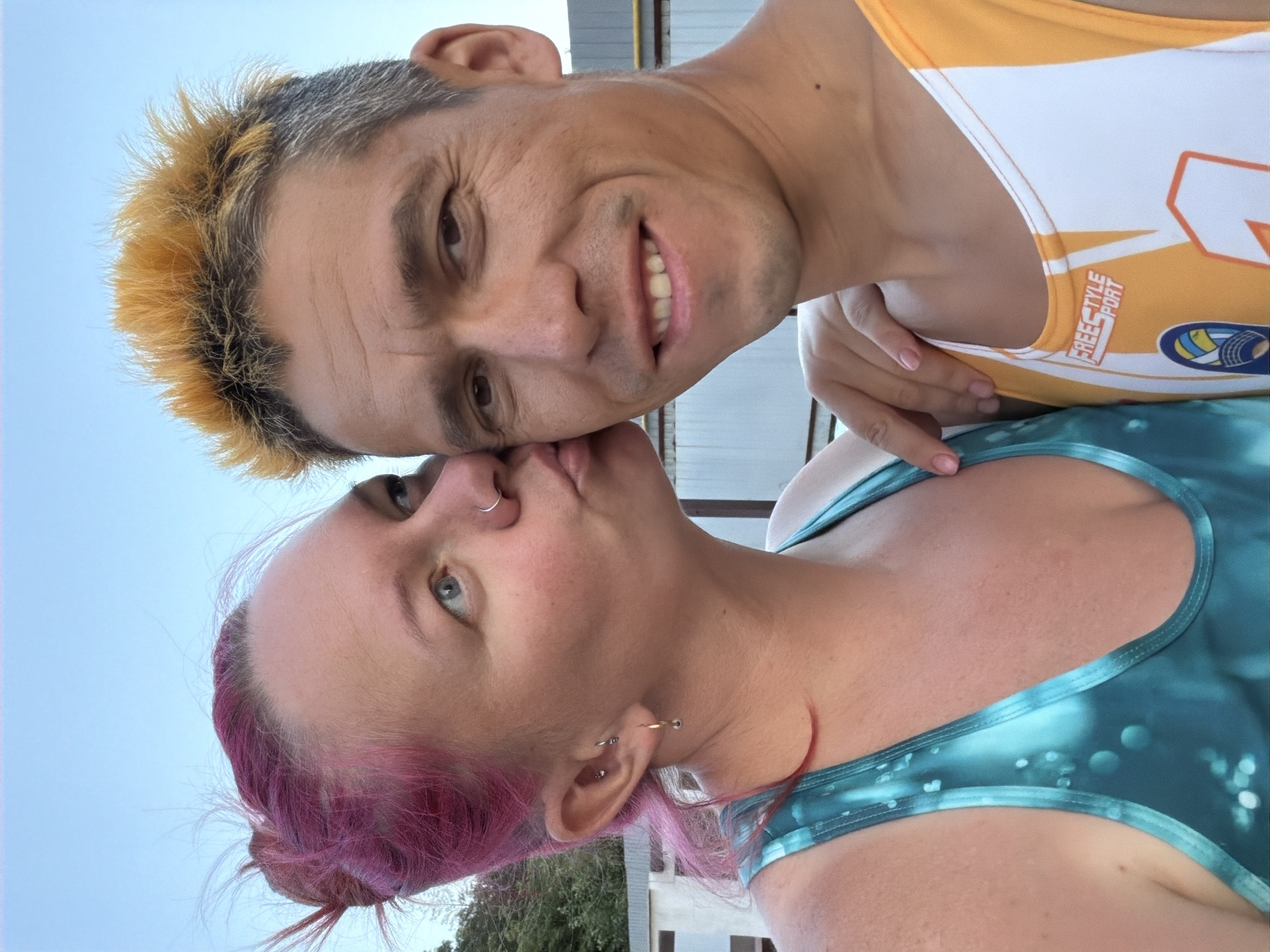 In a medium shot, a couple poses for a selfie outdoors against a light blue sky and a blurred background of buildings and trees. The woman, on the left, with bright pink hair styled in a messy bun, kisses the man on the cheek. She wears a turquoise and green tie-dye athletic tank top and has a hoop earring in her left ear and a nose ring. The man, on the right, has spiky, dyed blonde hair and a slight stubble. He is wearing an orange and white athletic tank top with the words "FREESTYLE SPORT" printed on it and a logo featuring a volleyball. His right arm is around the woman's shoulders, and his left hand is on her upper arm. The overall impression is of a candid and affectionate moment.