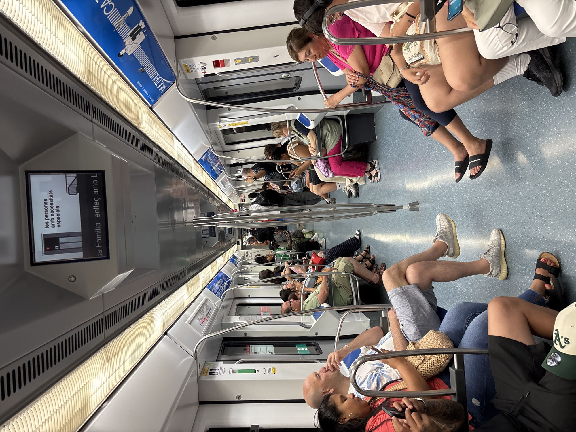 The image shows the interior of a Barcelona Metro train car, specifically on the L10 Sud line, as indicated by signage visible in the image. The train is populated with passengers seated and standing, illuminated by the overhead fluorescent lighting. The seats are arranged along the sides of the carriage, with metal handrails extending throughout the car for standing passengers. Passengers are dressed in casual summer clothing, suggesting warm weather. Some individuals are looking at their phones, while others are engaged in conversation or observing their surroundings. The environment is clean and modern, typical of a public transportation system. Advertising posters are visible on the upper walls of the train car.
