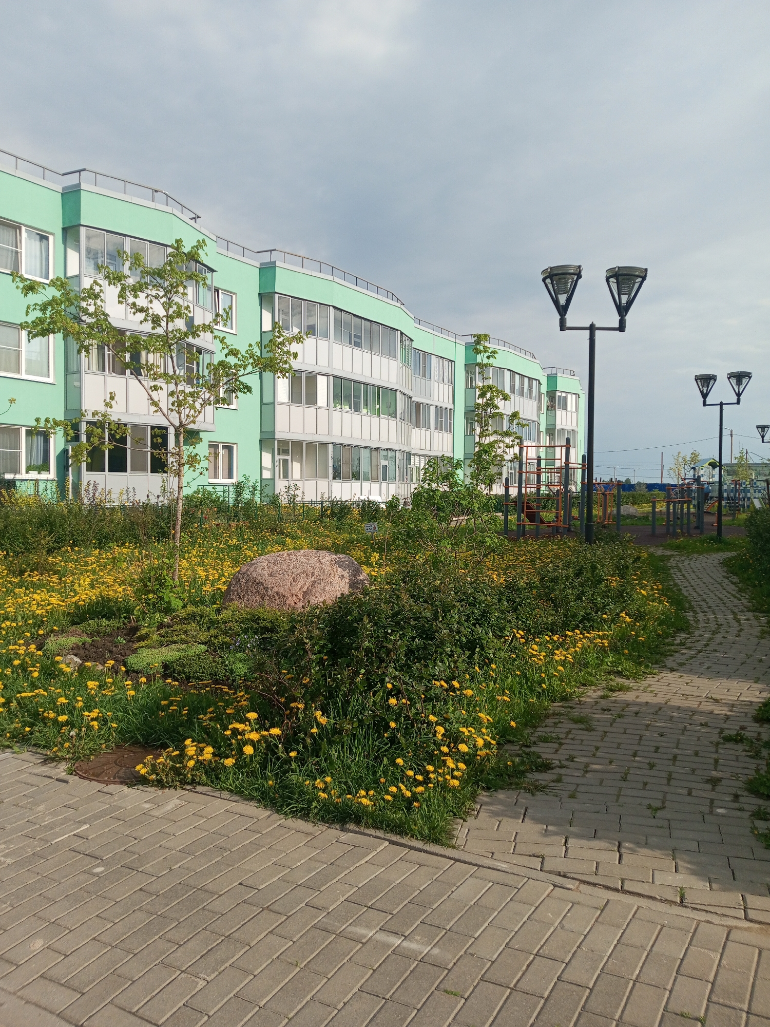 The image depicts an outdoor scene in Kommunar, Russia. A multi-story building with a pale green facade dominates the background. In the foreground, a brick pathway leads into a garden area filled with yellow dandelions and green shrubs. A large boulder sits within the garden. Several tall, black lampposts line the pathway. A playground is visible in the background. The sky is overcast with gray clouds.
