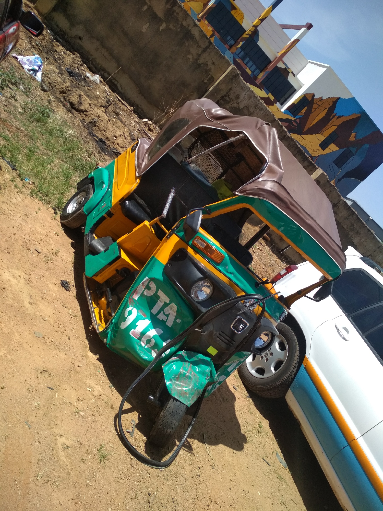 A three-wheeled vehicle (tuk-tuk) with green, yellow, and brown coloring is parked on a dirt and grass surface in the city of Unknown, Unknown. The vehicle's front is green with "PTA-916" visible. The rear section transitions to yellow and then brown on the top. Next to it is a white car with a blue and yellow stripe along the lower part of the door. In the background, there's a concrete wall and a building with a mural. The sky is visible in the upper right.