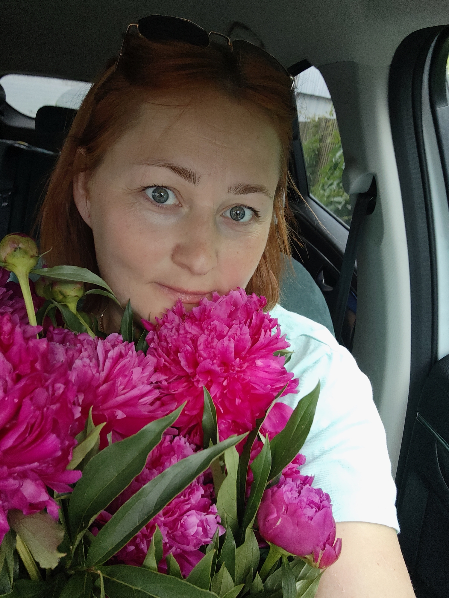 A woman with auburn hair and blue eyes is in the driver's seat of a car, holding a bouquet of bright pink peonies. She is looking directly at the camera with a slight smile. Sunglasses are perched on top of her head. The interior of the car is visible, including the steering wheel and part of the dashboard. Outside the car window, lush green foliage and a wooden fence can be seen, suggesting a location with natural surroundings. The scene appears to be in a suburban or rural area of Noviy, Russia.