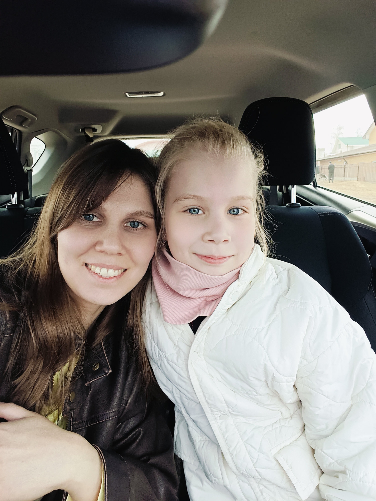 The image displays a close-up, eye-level portrait of two females, an adult woman and a young girl, sitting side-by-side in the back seat of a car. Both subjects are looking towards the camera with gentle smiles, suggesting they are posing for a photograph.
The woman, on the left, has long brown hair that frames her face and distinct blue eyes. She is wearing a dark brown, leather-textured jacket over a yellow top, and her smile is broad, revealing her teeth.
Beside her, on the right, is a young girl with blonde hair and bright blue eyes. She has a soft, subtle smile. She is dressed in a light pink turtleneck or scarf and a white quilted puffer jacket.
The car's interior is partially visible, showing dark fabric seats, headrests, and the light-colored ceiling with an overhead light fixture.
Through the car windows on the right, an outdoor scene is visible, suggesting a daytime setting with diffused light, possibly indicating overcast weather. The background shows a wooden fence, distant houses with light-colored roofs (one appears green), and bare trees. Two small, indistinct figures, appearing to be men, can be faintly seen standing beyond the fence.
The overall mood conveyed by the subjects' expressions is pleasant and content. No visible text is present in the image.