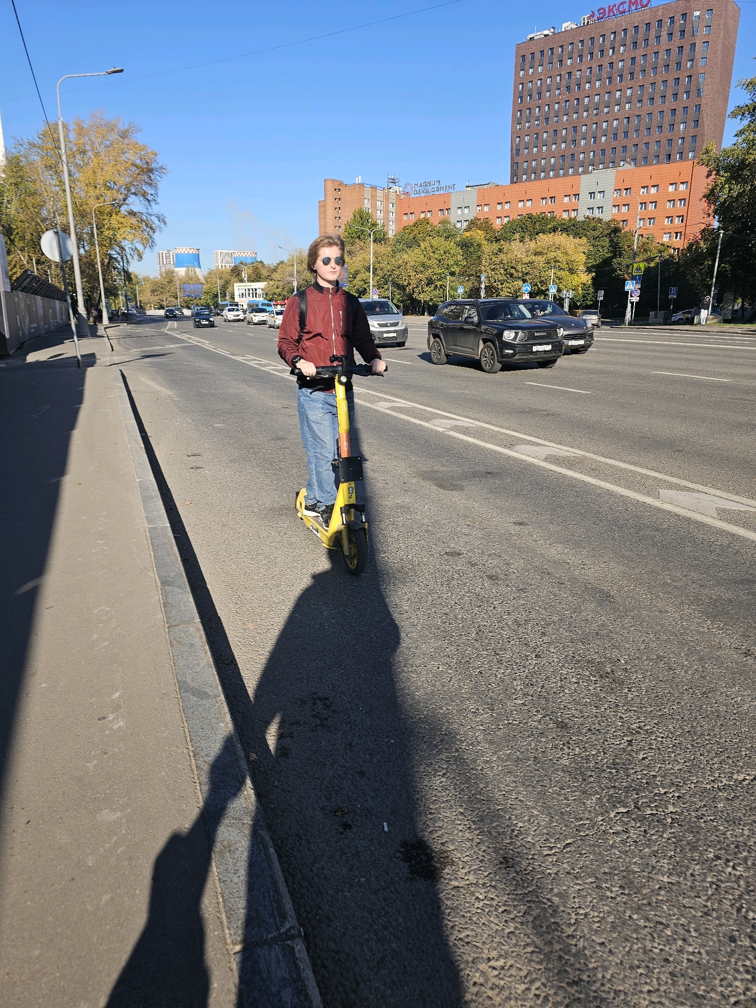 A young person with blonde hair, wearing sunglasses, a maroon jacket, and blue jeans, rides a yellow electric scooter on an asphalt road. The person is looking forward, holding the scooter's handlebars. A long shadow of the person and scooter is cast on the road and sidewalk. In the background, there is a multi-story brick building with the red inscription "ЭКСМО" at the top. Further in the background, there are more buildings, trees with yellowing leaves, and a clear blue sky. Cars are visible on the road. The scene appears to be a street in Moscow, Russia, on a sunny day.