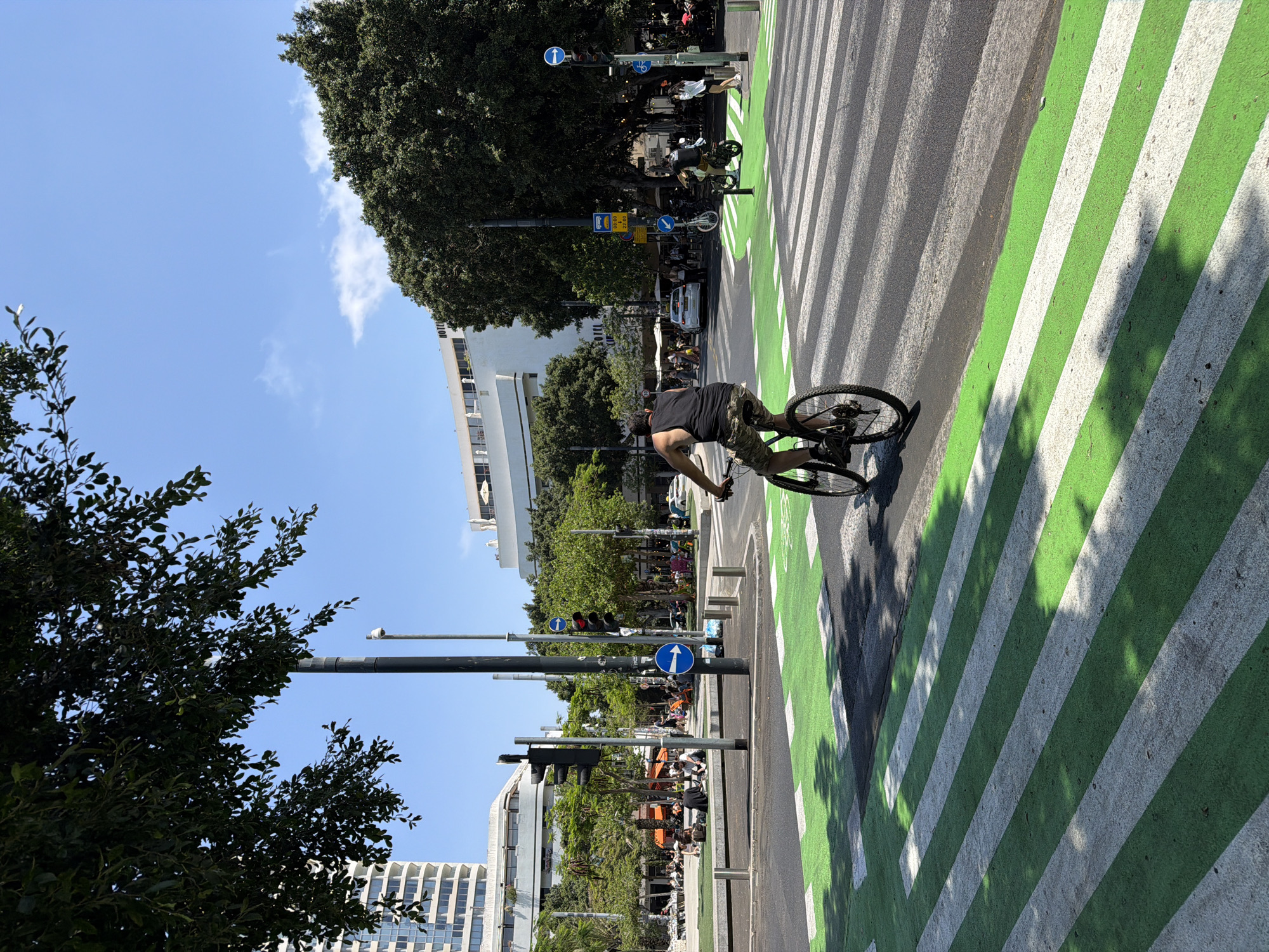 The image captures a street scene in Tel Aviv, Israel. A man on a bicycle is riding across a green and white striped bike lane. There are trees, lampposts, traffic lights, and street signs visible along the sidewalk. Several buildings are visible in the background, including a modern white structure. The sky is blue with a few scattered clouds. People are visible sitting outside at tables in the background.