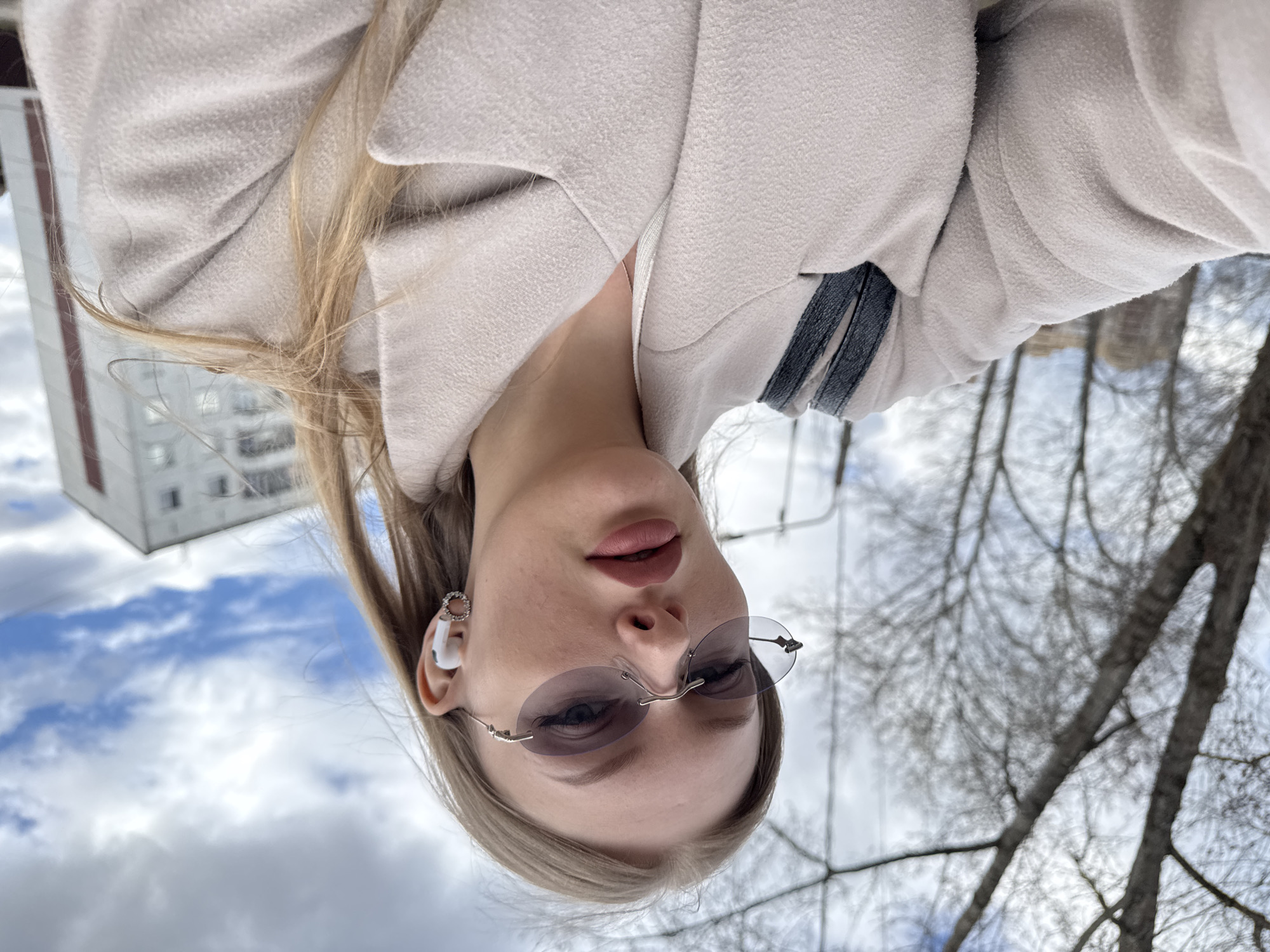 This image is a close-up portrait, oriented upside down, featuring a young woman against an outdoor backdrop. The woman has blonde or light brown hair and is wearing a light beige or off-white coat. She sports small, rimless sunglasses with lightly tinted lenses. A white earbud is visible in her right ear, accompanied by a small, intricate circular earring. Her lips are slightly parted, adorned with light pink lipstick.
The background reveals a cloudy sky, leafless tree branches, and a distant white building with visible windows, suggesting an urban or suburban setting. The bare trees indicate that the photograph was taken during late fall, winter, or early spring. The overall lighting suggests it is daytime, under overcast conditions. The subject appears to be looking directly at the camera, implying a selfie or a directed portrait. There is no visible text in the image. The location context provided is Perm, Russia.