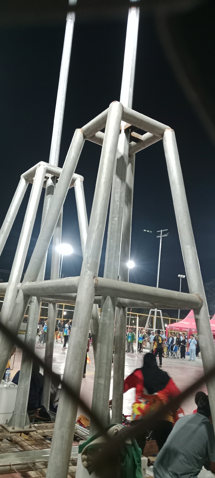 The image depicts a night scene at the Youth Center in Tuban, Indonesia, likely during an event or gathering. Dominating the foreground is a large, industrial-looking metal structure, possibly part of a stage or lighting rig, made of thick, hollow cylindrical pipes. The structure features a cubic framework at the top, with angled supports extending downwards. The scene is illuminated by bright floodlights positioned above, casting harsh shadows.
In the background, several people are visible, some in athletic wear and others in casual attire. They appear to be engaged in activities on a paved outdoor court. There are also red tents set up, suggesting vendor stalls or event spaces. A person in a black hijab and a brightly colored outfit is in motion in the mid-ground. Another person wearing a grey shirt is in the lower right corner. The overall environment suggests a public event or sports competition taking place after dark.