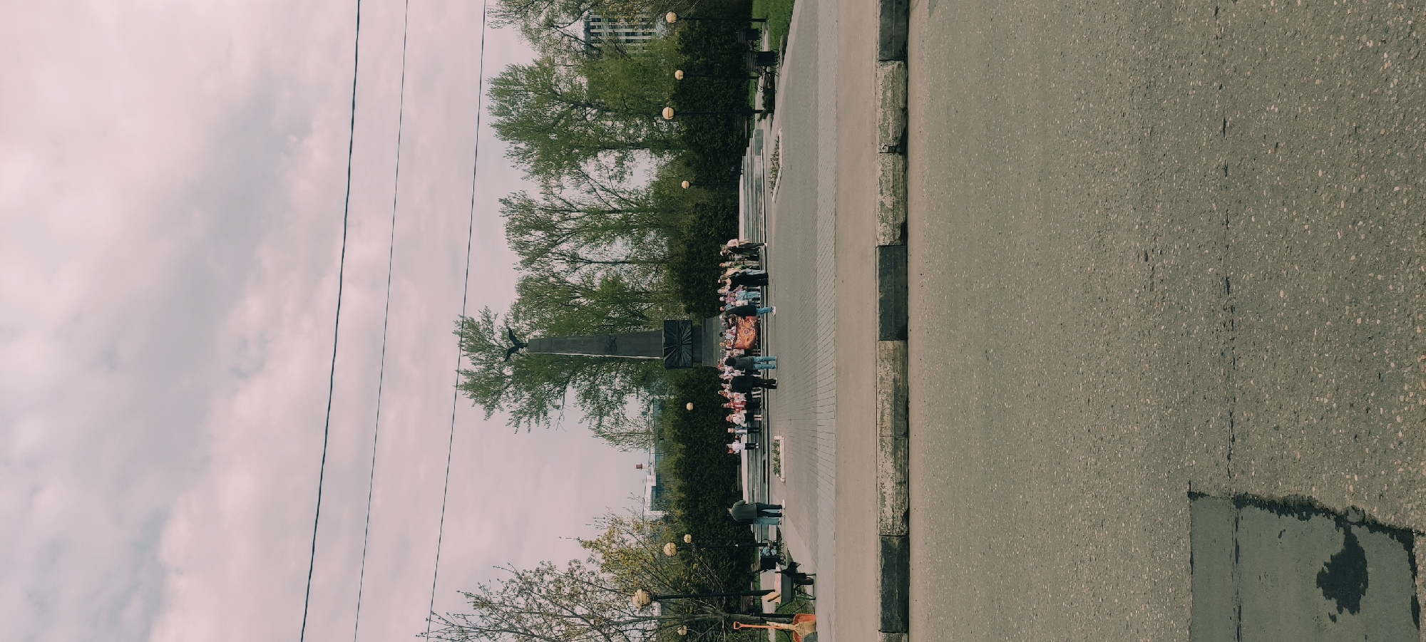 A photograph of a monument in Fryazino, Russia, taken on an overcast day. The monument, featuring an eagle atop a pillar and a star symbol, stands amidst greenery and trees. A gathering of people, including adults and children, is present at the monument. The image includes overhead power lines and a paved road with a concrete curb on the right side. A figure stands near a bench and an orange shovel on the bottom left of the image.